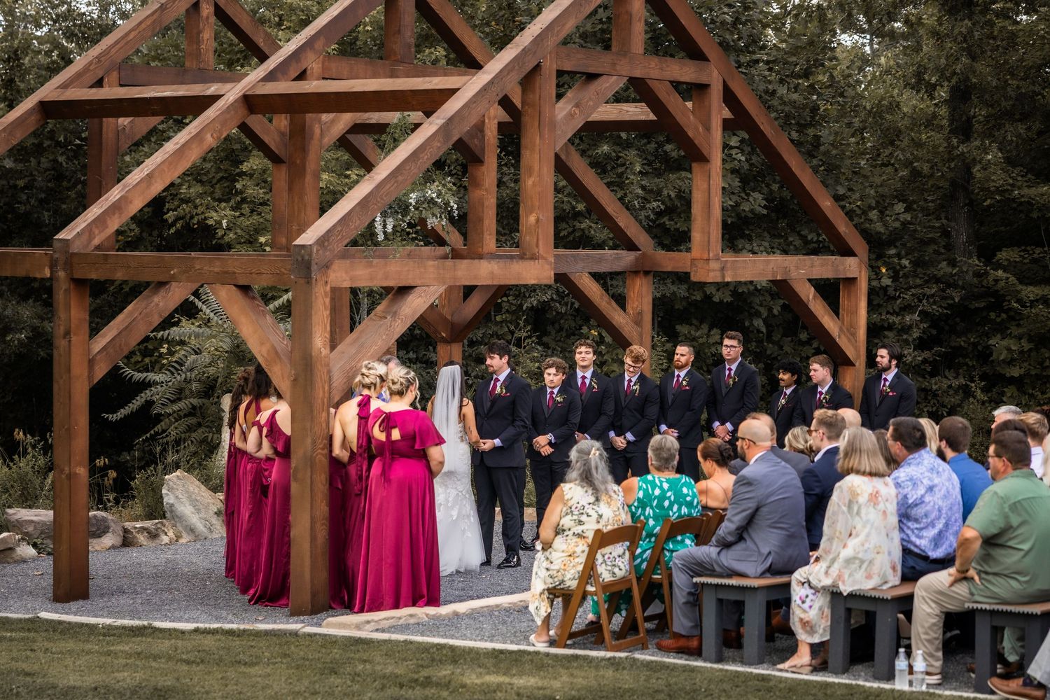 Outdoor wedding ceremony under a rustic wooden timber frame arch, with bridesmaids in burgundy dresses and guests seated on benches.