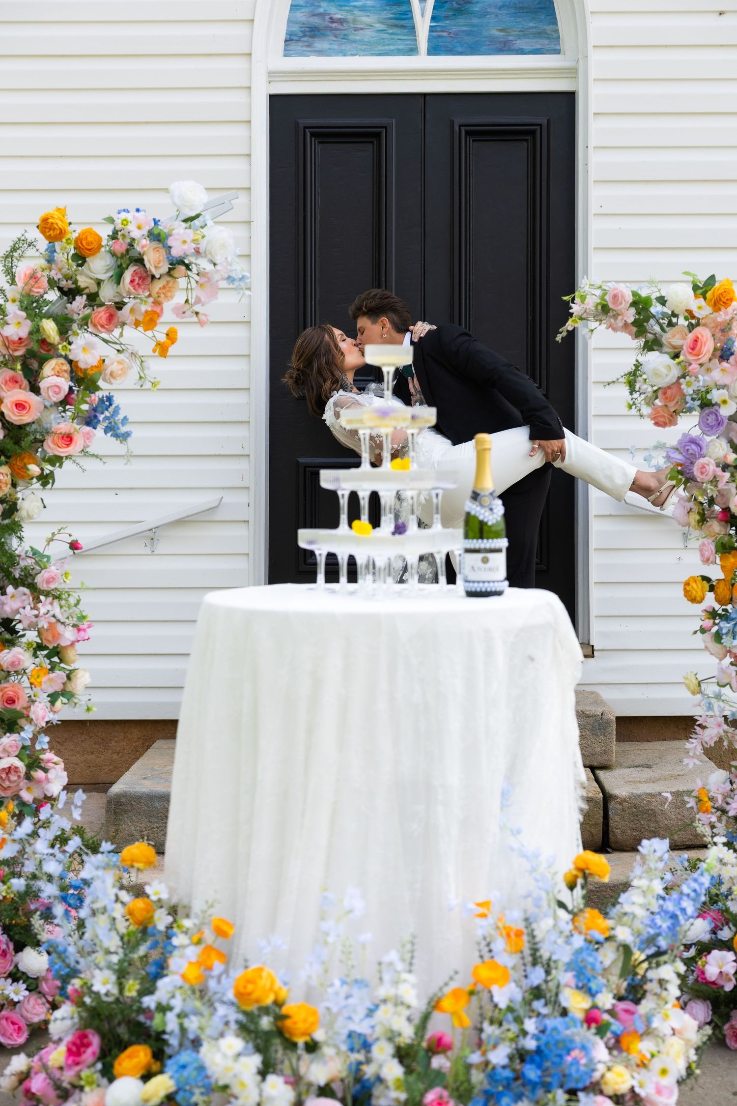 Newlyweds sharing romantic dip kiss beside champagne tower with colorful floral arch.