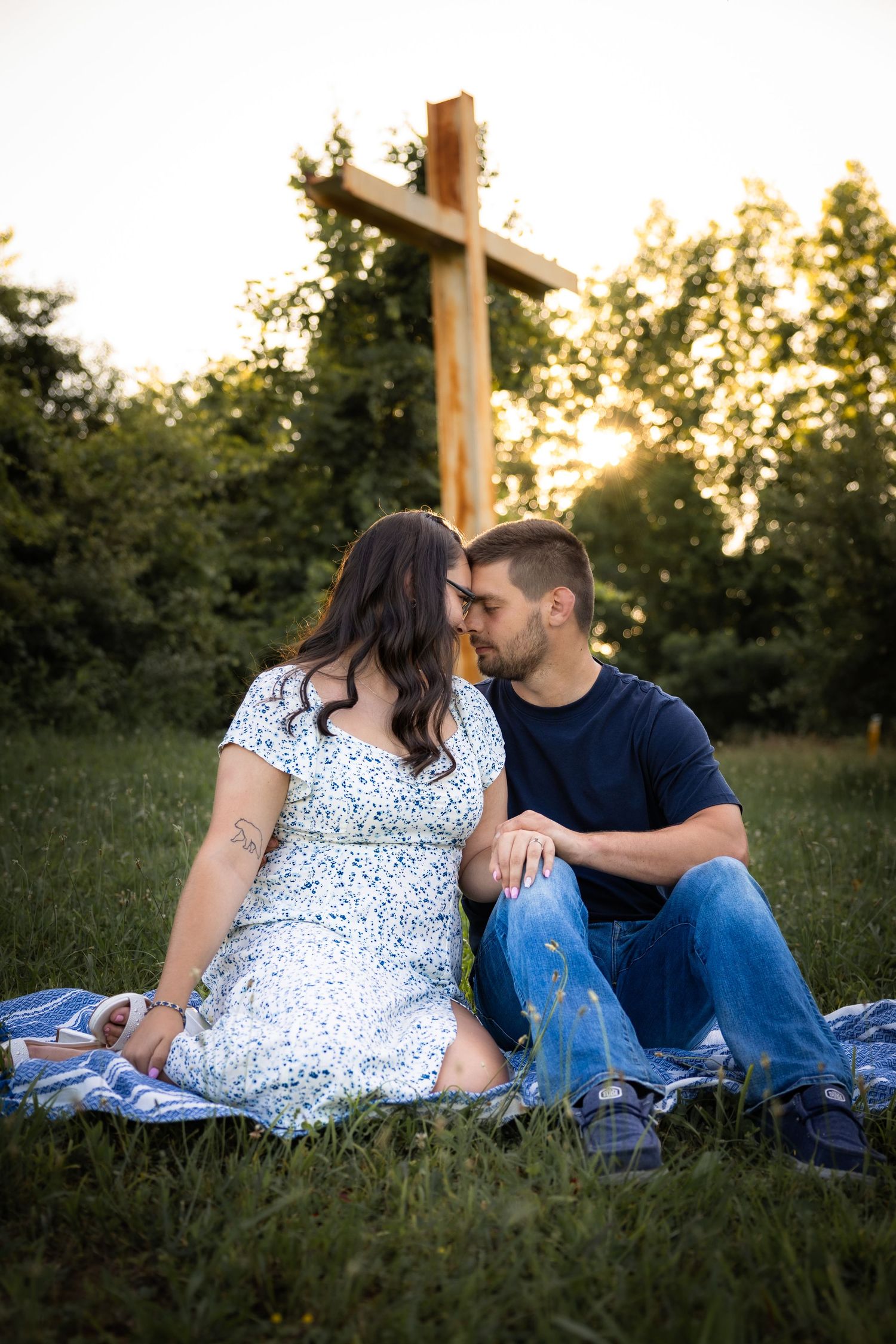 Couple sharing a romantic moment on a blanket in a grassy field with a wooden cross and golden sunset behind them.