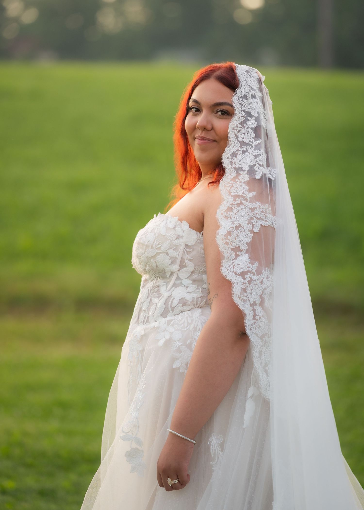 Bride in lace wedding gown and veil with red hair posing outdoors on green grass at golden hour.