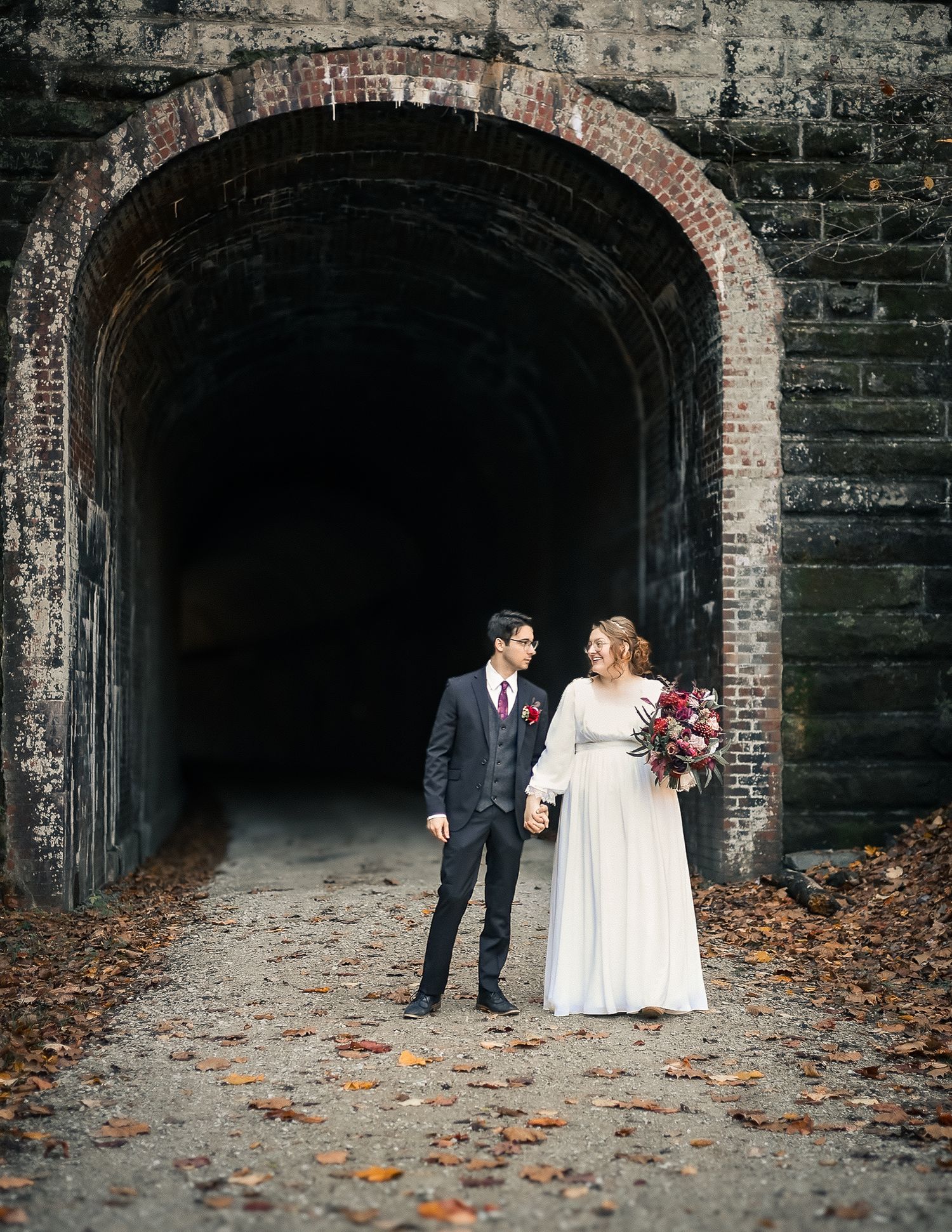 Newlywed couple holding hands and gazing at each other inside a rustic brick tunnel archway.