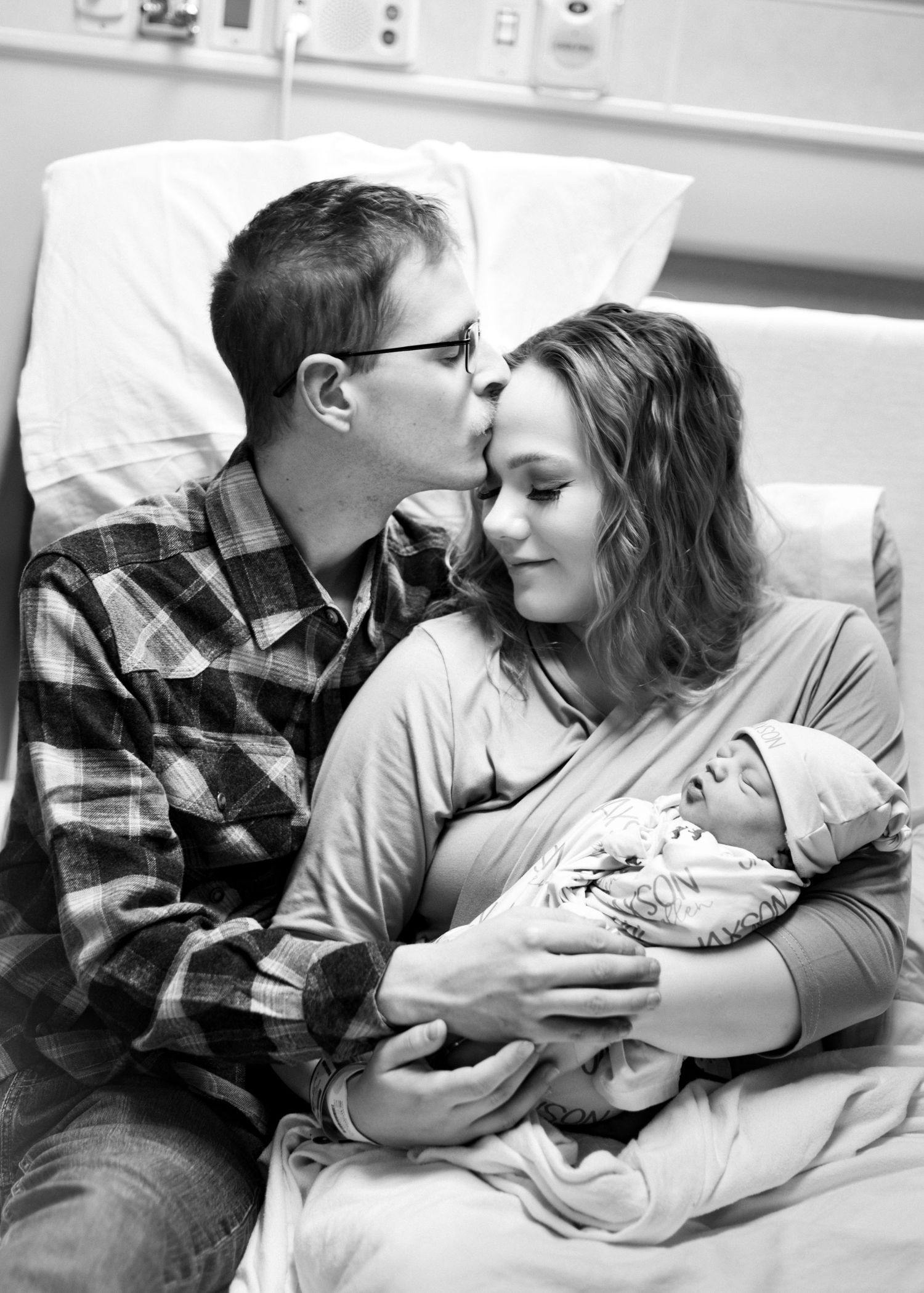 New father kissing mother on forehead as she holds newborn baby in hospital bed, black and white photo.