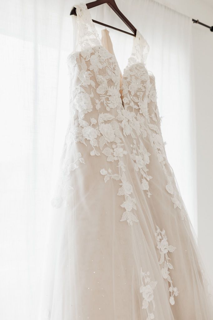 Elegant white lace applique wedding gown hanging on wooden hanger against bright window.