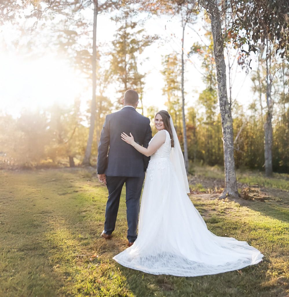 Bride and groom walking in golden sunset light among tall trees in wedding attire.