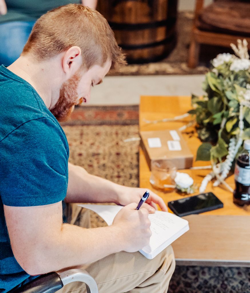 Red-haired groom in teal shirt writes heartfelt vows in a notebook at a rustic wedding venue.