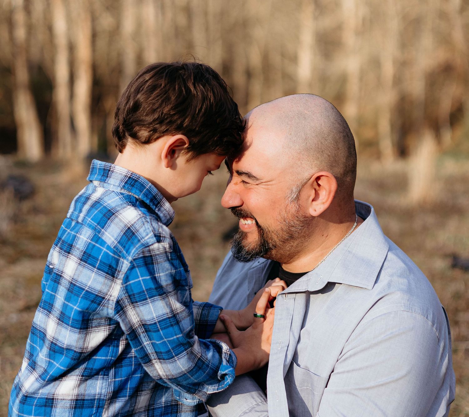 Loving bald father touching foreheads with young son outdoors in sunny forest setting.