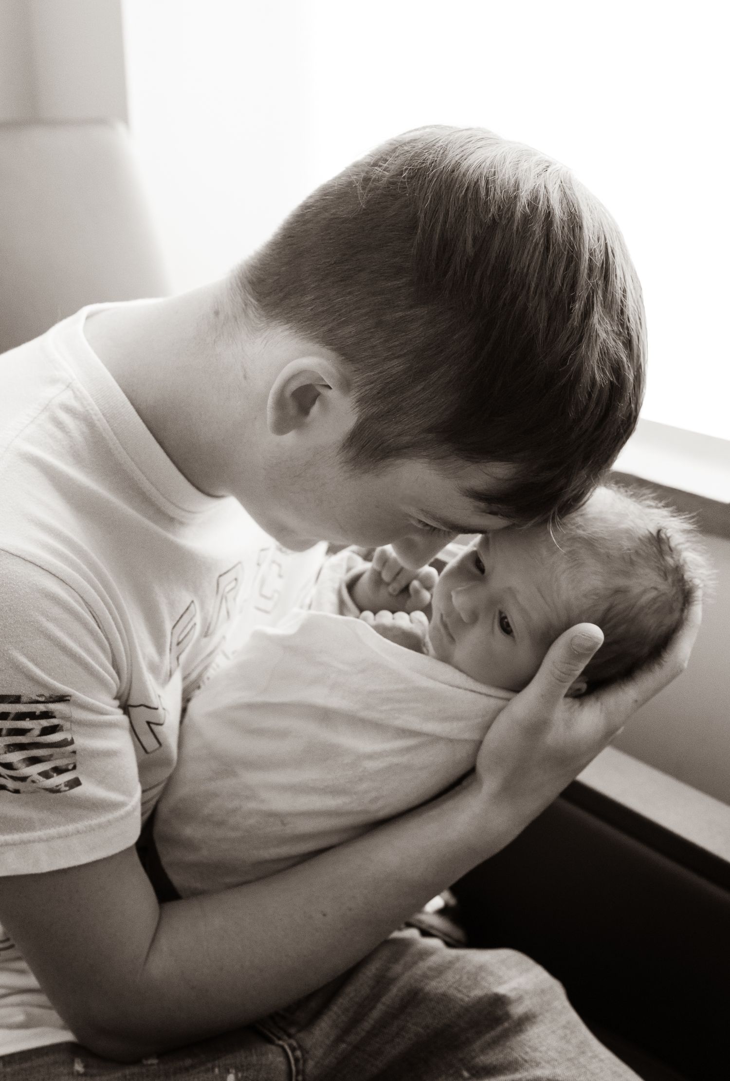Black and white photo of a young father gazing lovingly at his newborn baby by window.