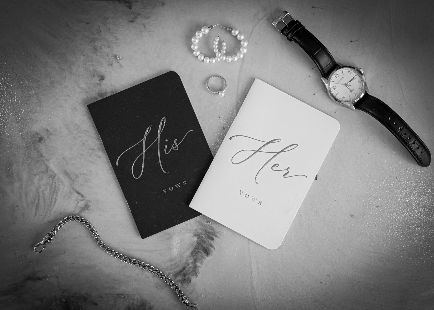 Black and white flat lay of His and Her vow books with a watch, pearl bracelet, and rings on marble.