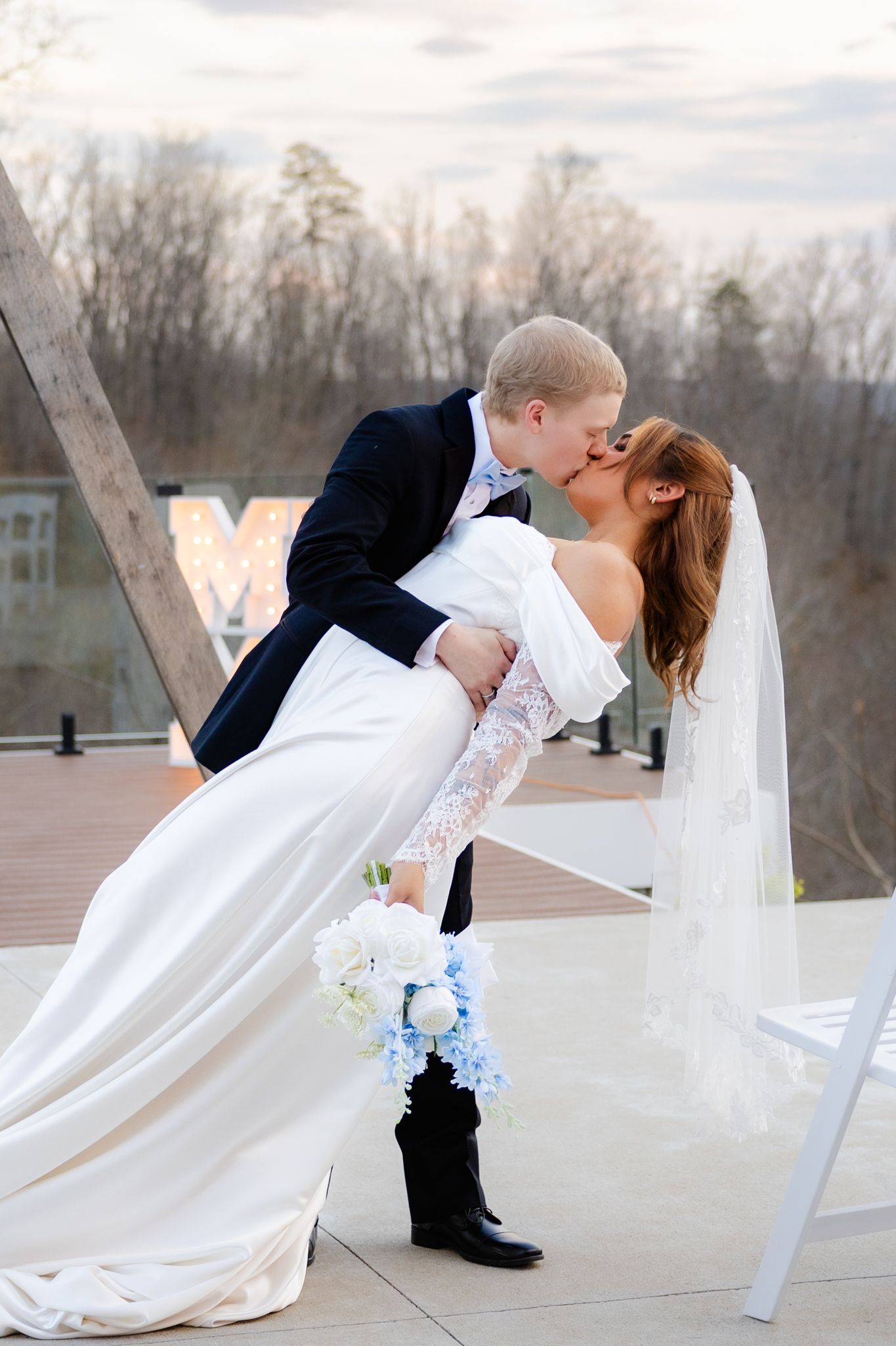 Groom dips bride for a passionate kiss outdoors, bride holding a blue and white bouquet in a lace wedding gown.