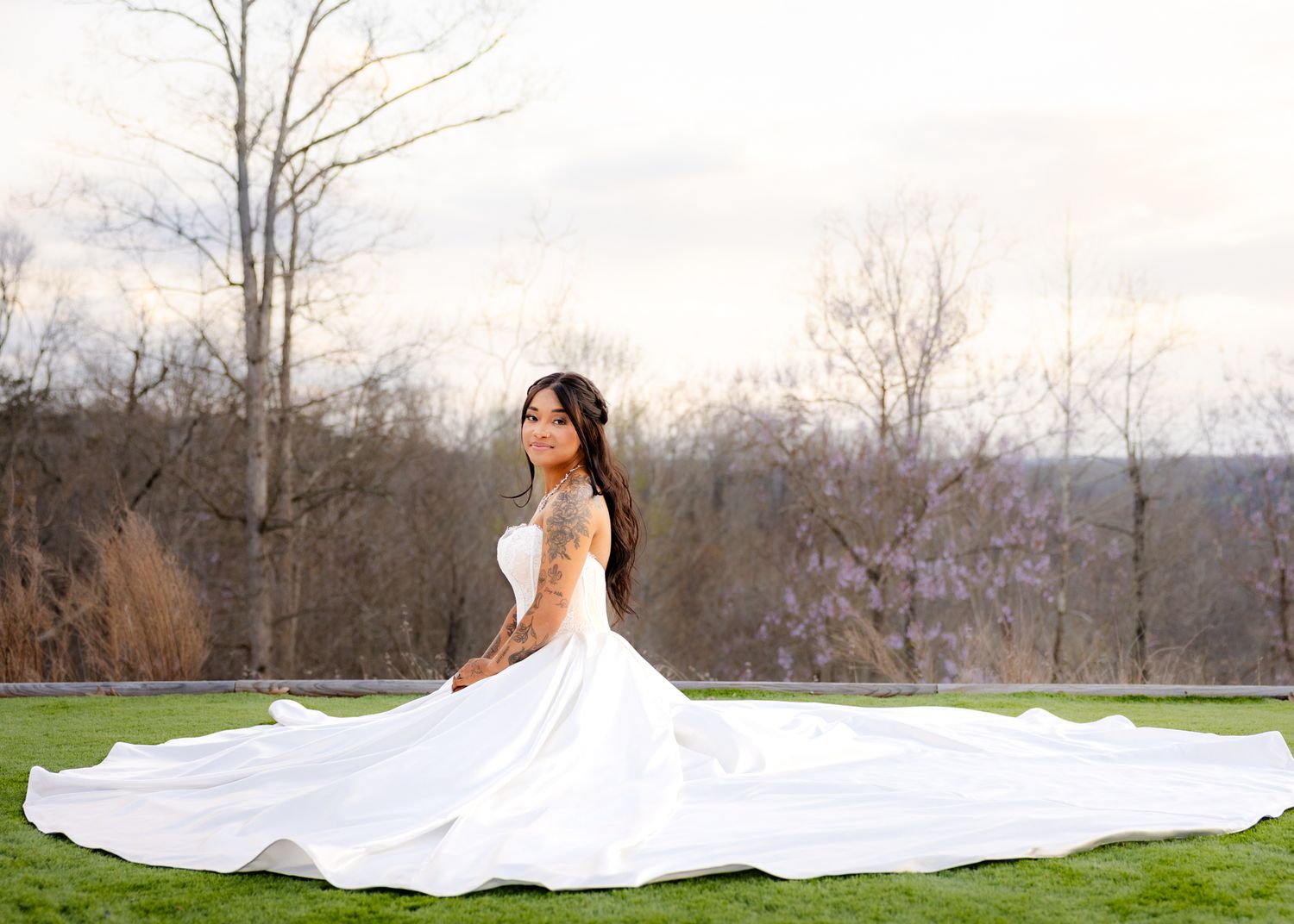 Bride in a stunning white ball gown sitting gracefully on green grass with trees in background.