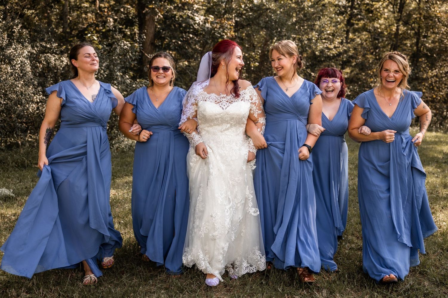 Bride in a white lace gown laughs while walking arm in arm with five bridesmaids in matching blue dresses on a grassy lawn.