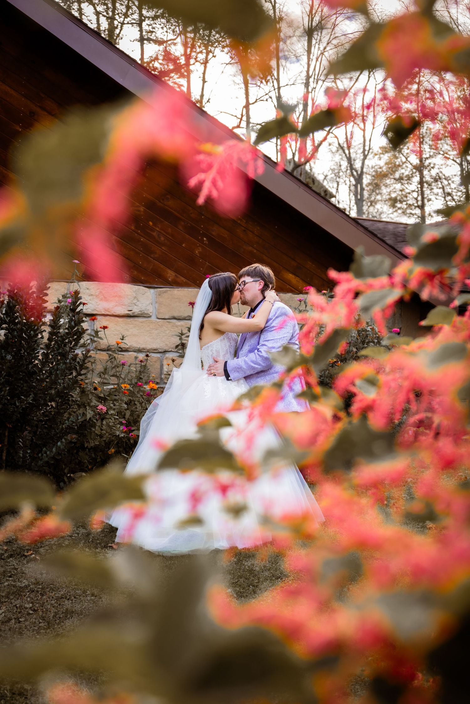 Bride and groom share a romantic kiss surrounded by vibrant red autumn foliage near a rustic wooden building.