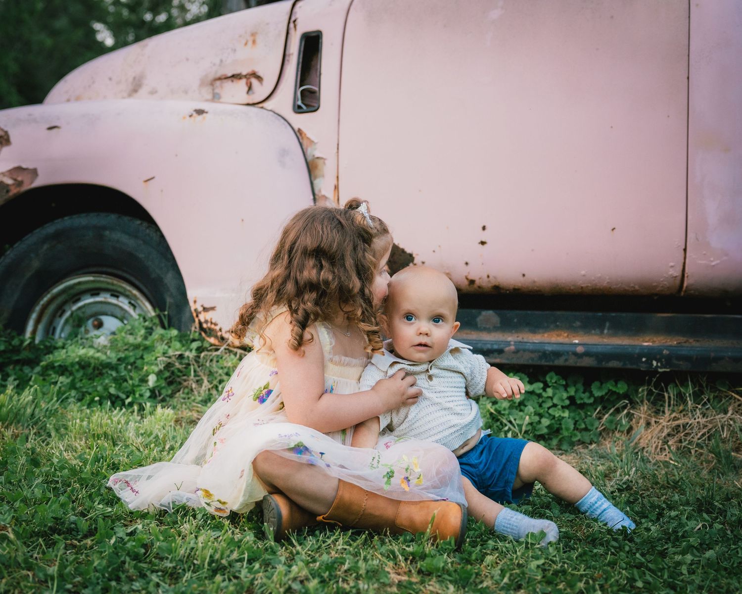 Young girl in floral dress kisses baby sibling on grass beside a vintage rusty pink truck outdoors.