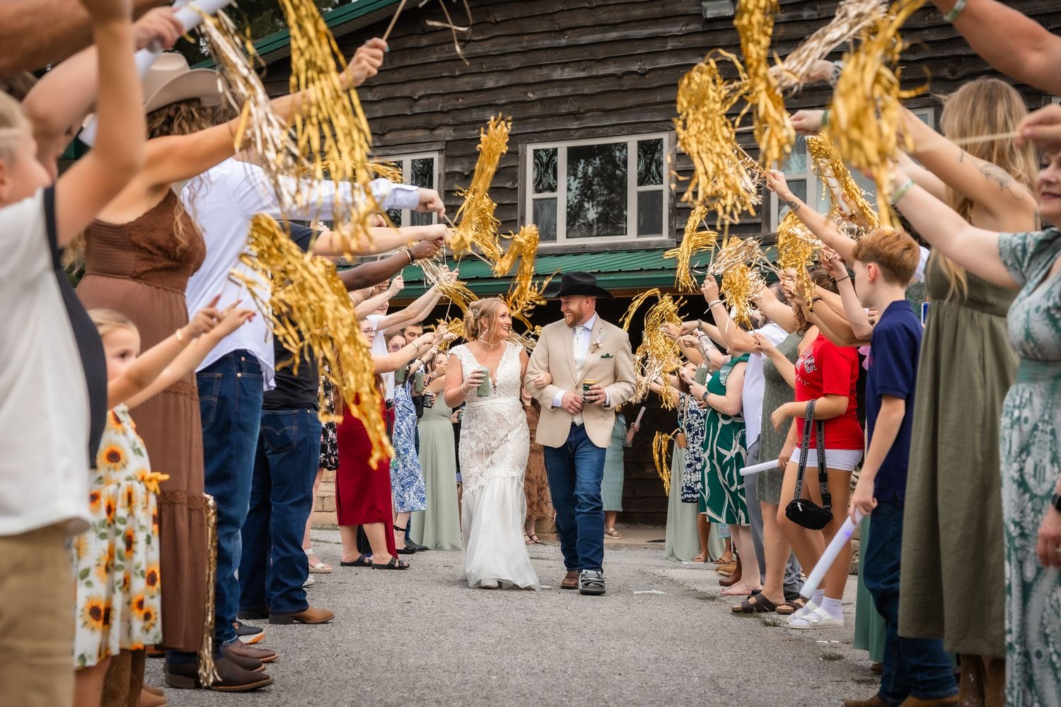 Bride and groom walk through wheat sheaf arch surrounded by cheering guests at rustic outdoor wedding ceremony.