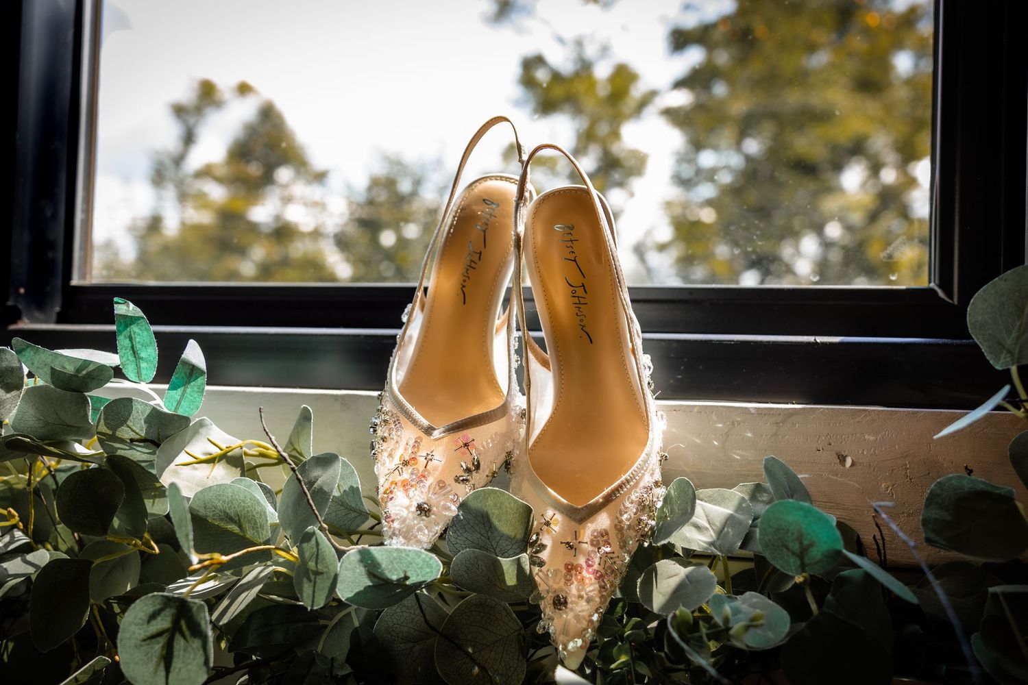 Elegant bridal heels with floral embellishments displayed on eucalyptus greenery in a sunlit window.