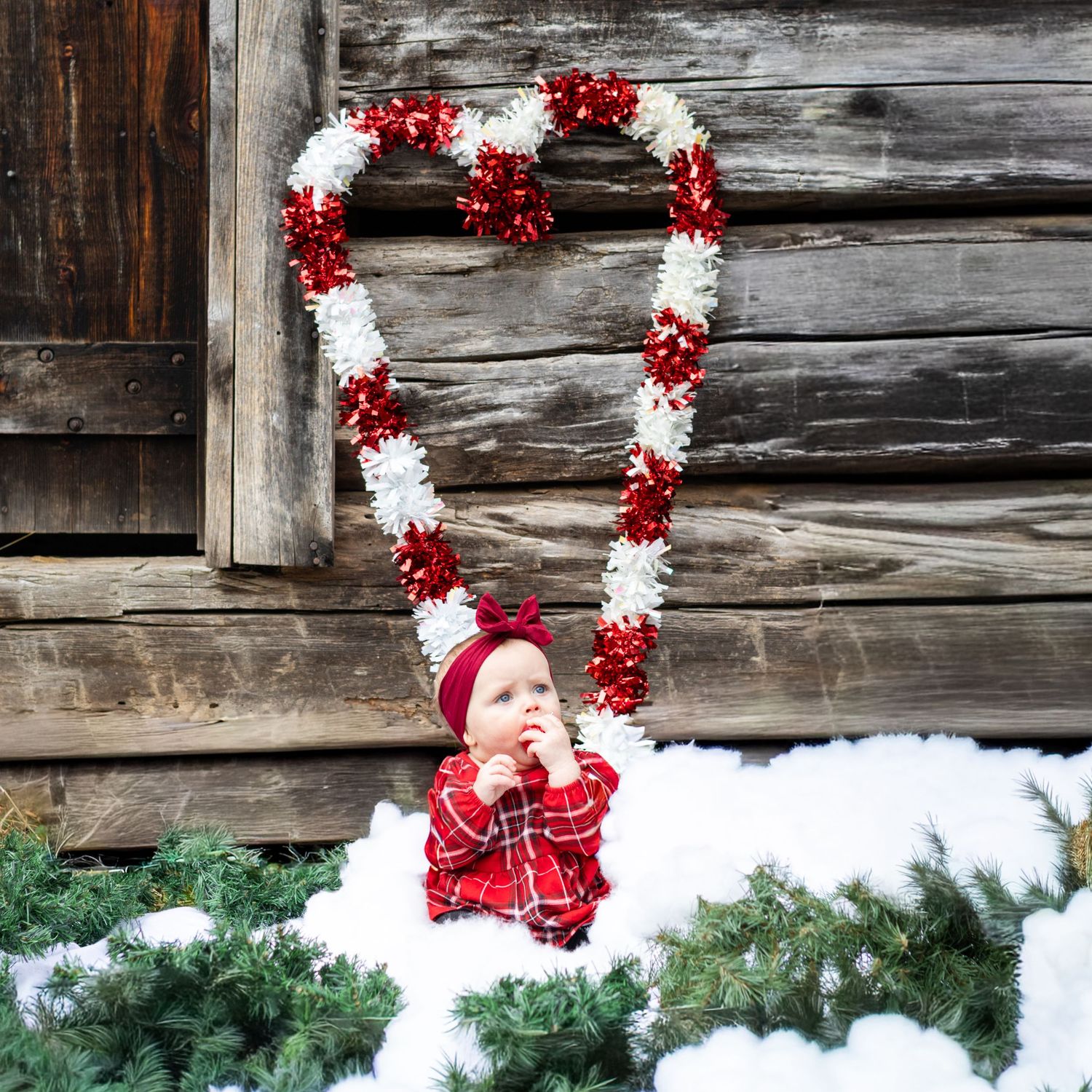 Baby in red plaid outfit sitting in snow inside a red and white heart-shaped Christmas wreath near a rustic wooden barn.