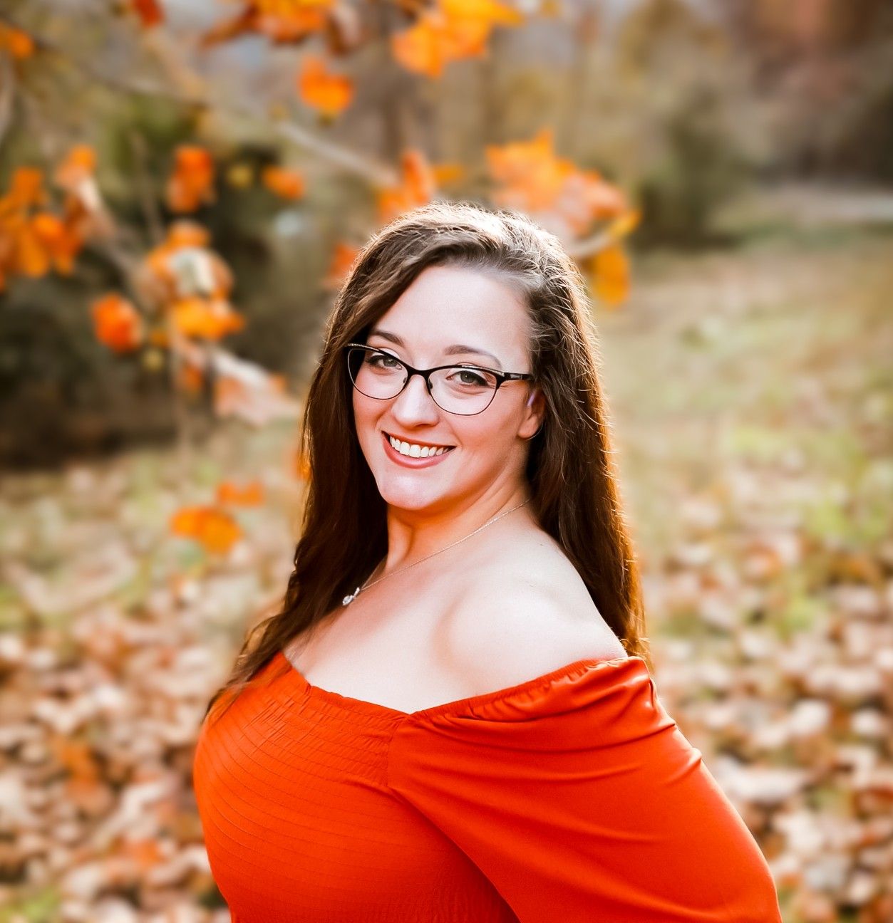 Smiling woman in an off-shoulder orange top poses among vibrant autumn leaves.