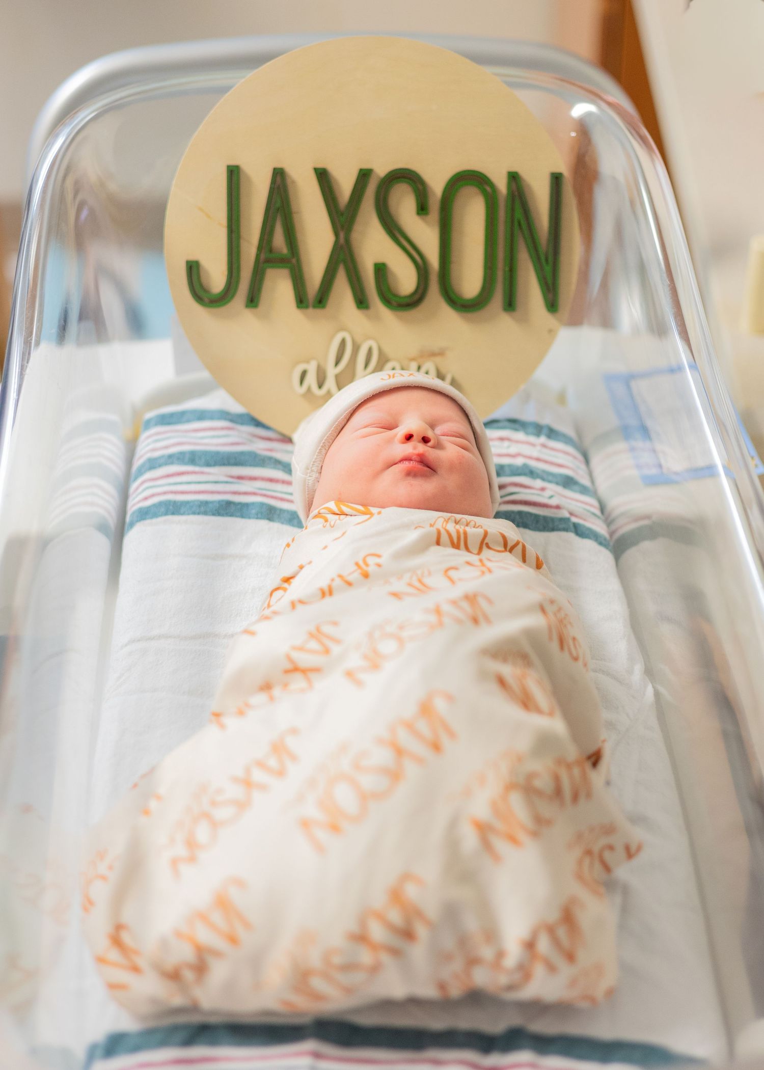 Newborn baby Jaxson swaddled in personalized blanket in hospital bassinet with name sign.
