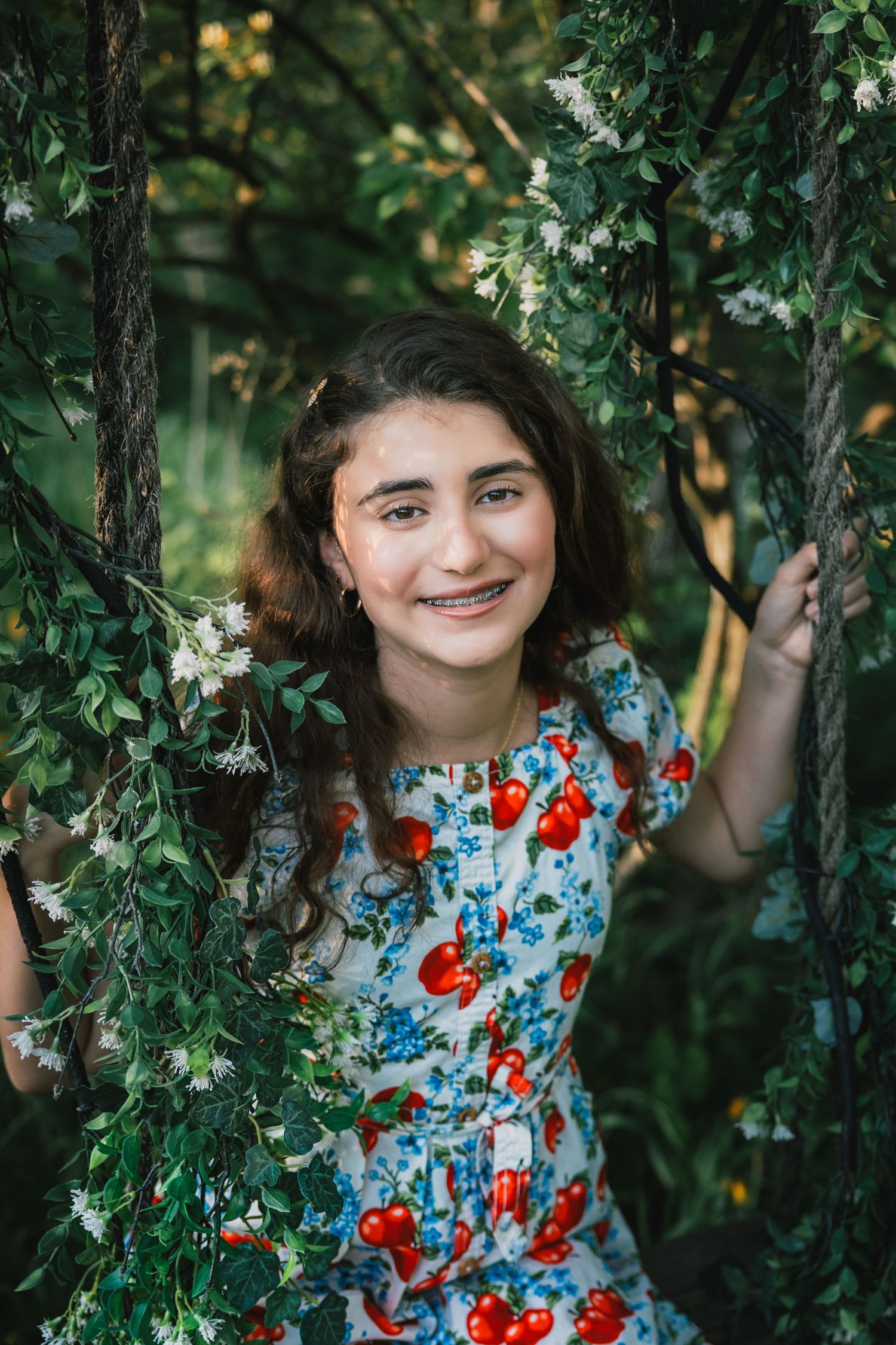 Smiling teenage girl with braces in cherry-print dress sitting on floral swing surrounded by green trees.
