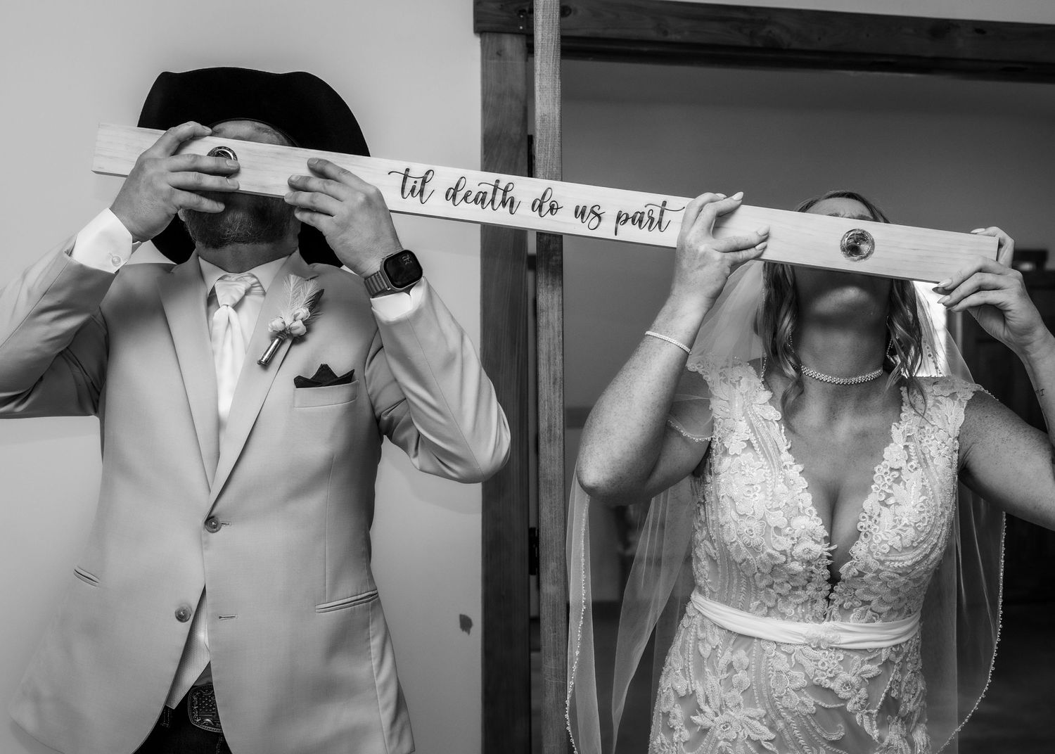 Black and white wedding photo of bride and groom holding a sign reading til death do us part covering their faces.