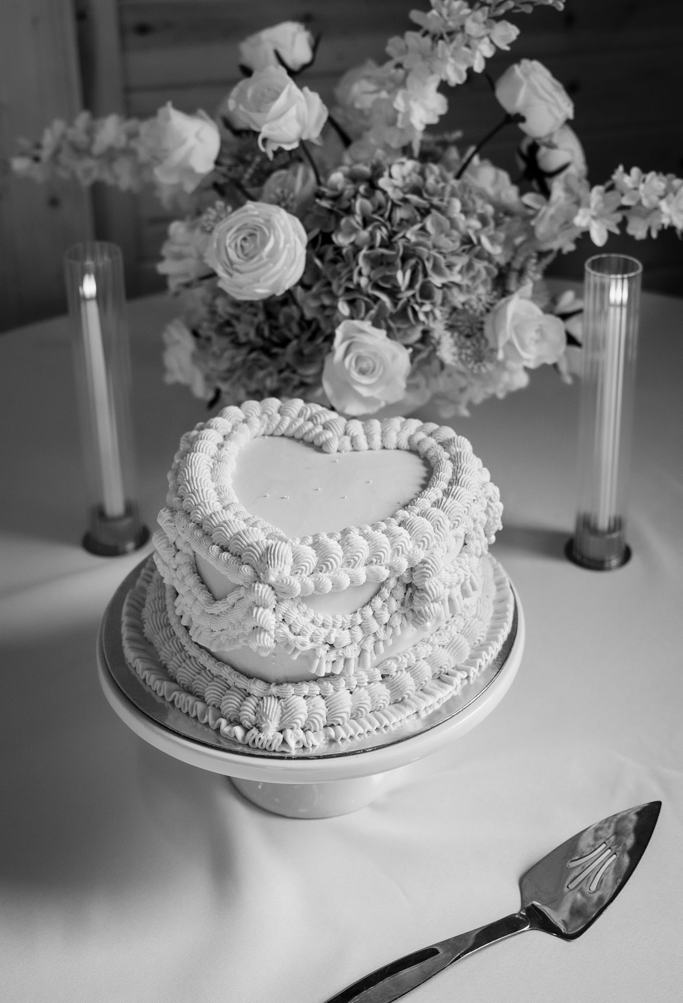 Black and white photo of heart-shaped wedding cake with roses and candles on table.