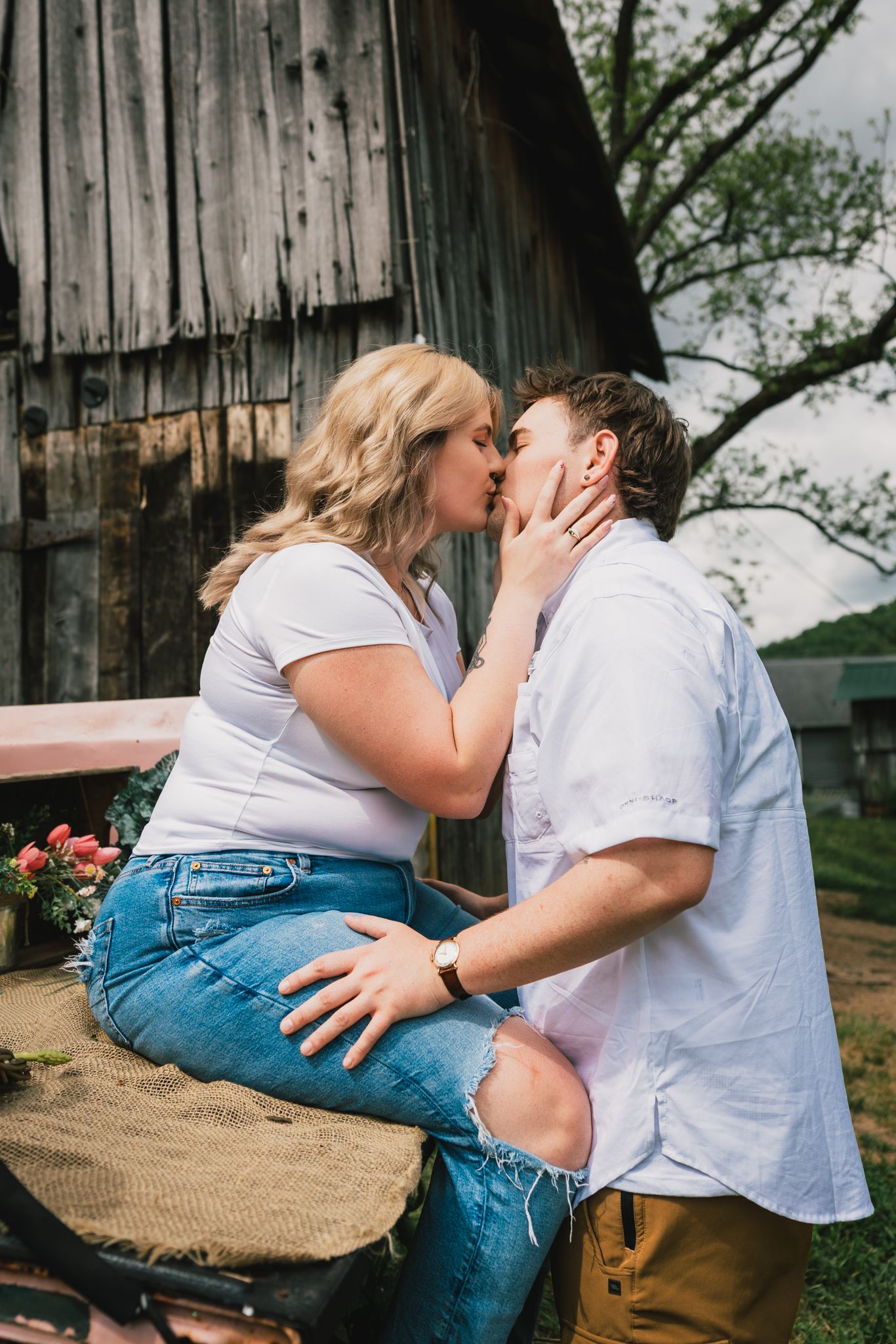 Young couple sharing a passionate kiss near a rustic barn with pink flowers and vintage tractor.