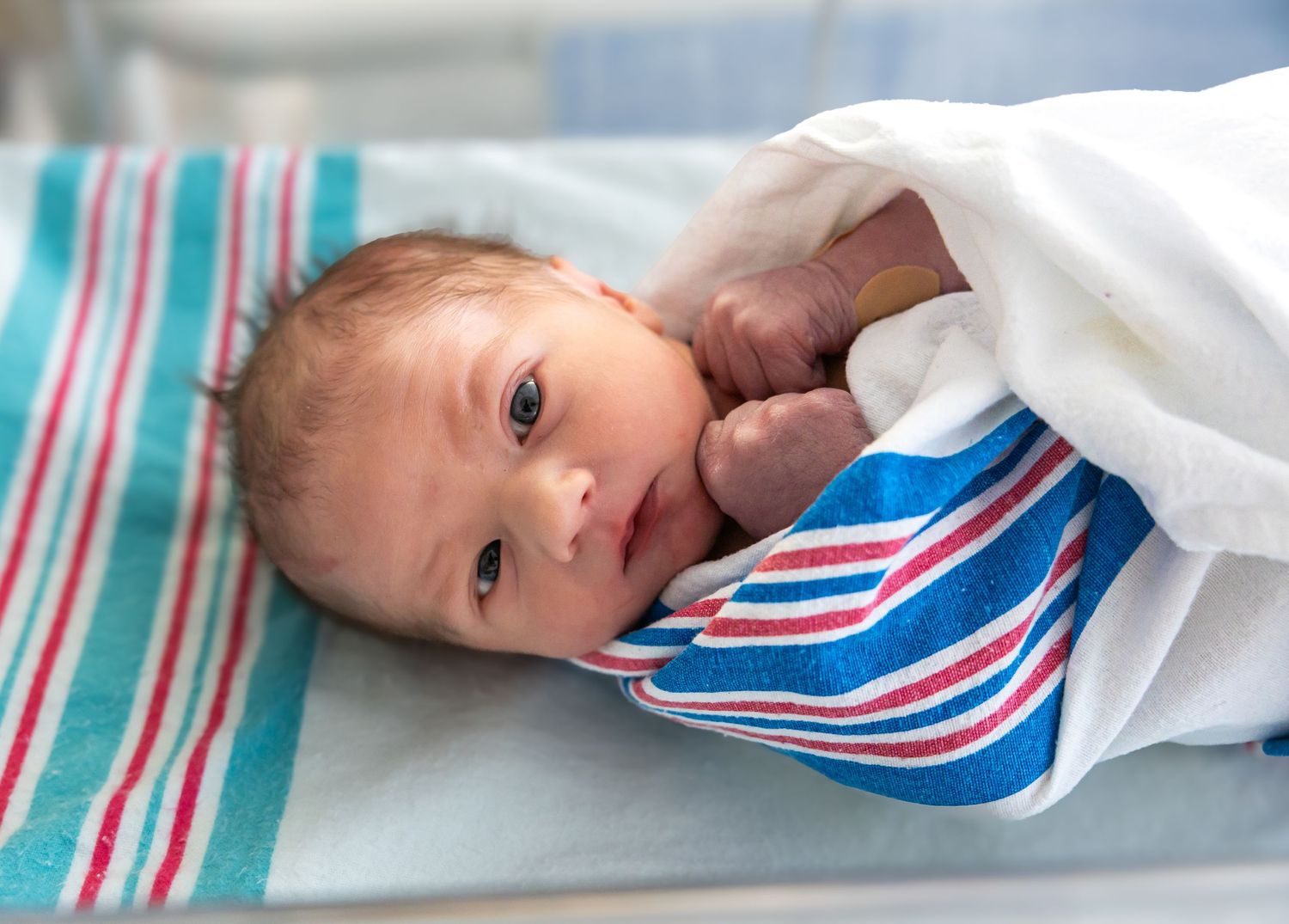 Newborn baby with dark hair lying awake in a striped hospital bassinet wrapped in blanket.