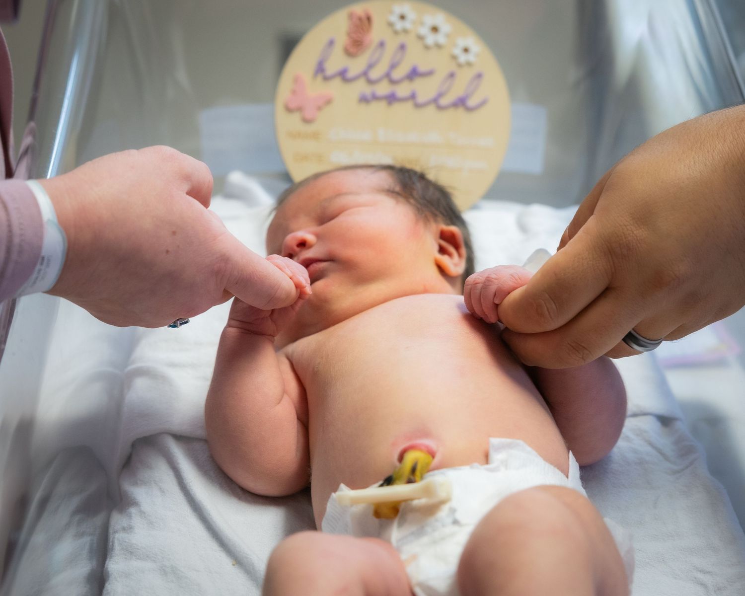 Newborn baby lying in bassinet with parents holding tiny hands next to a hello world sign.