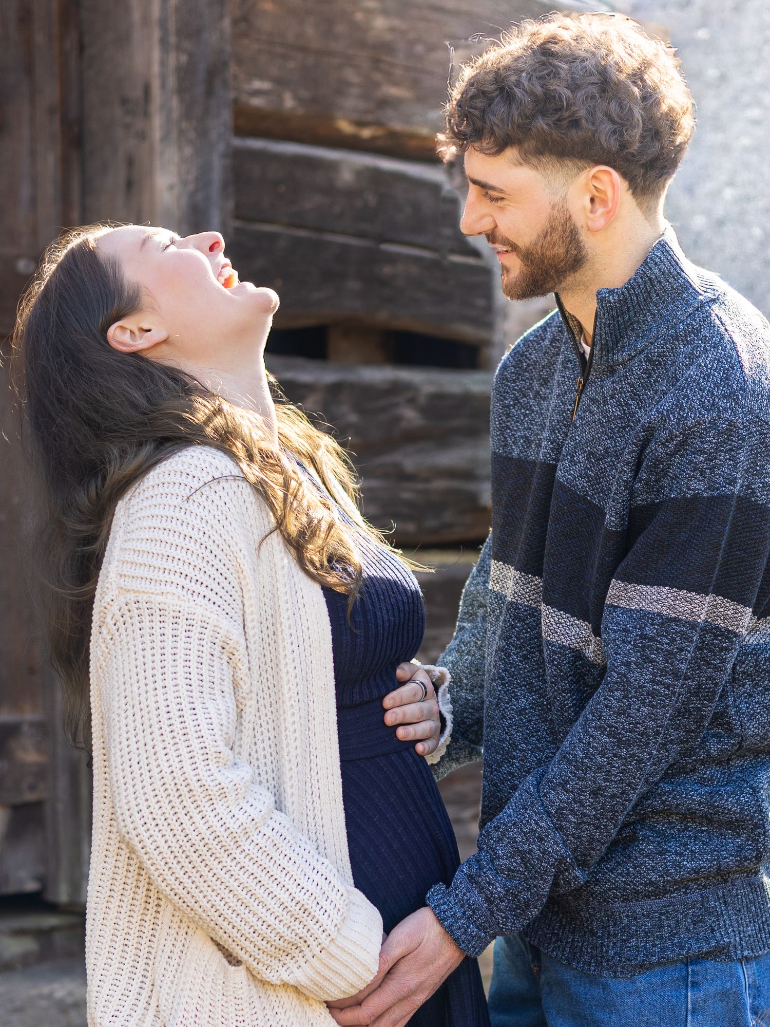 Happy expecting couple laughing together outdoors during a maternity photo session near rustic barn.