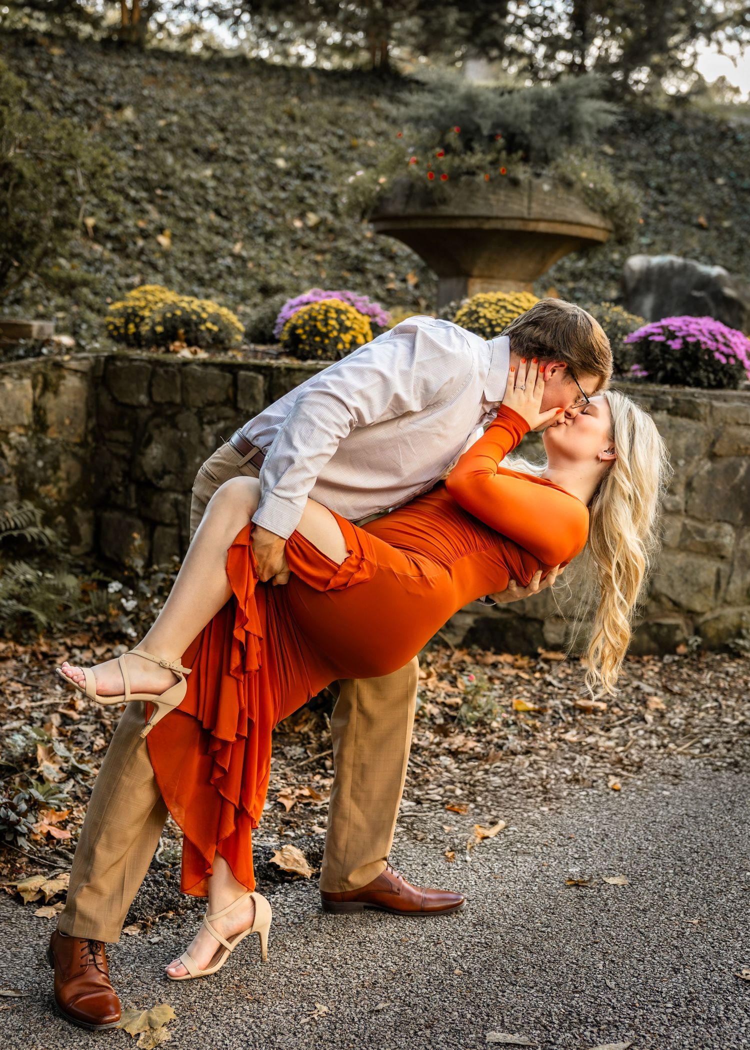 Couple sharing a romantic dip kiss outdoors in fall garden, woman in orange dress, man in white shirt and tan pants.