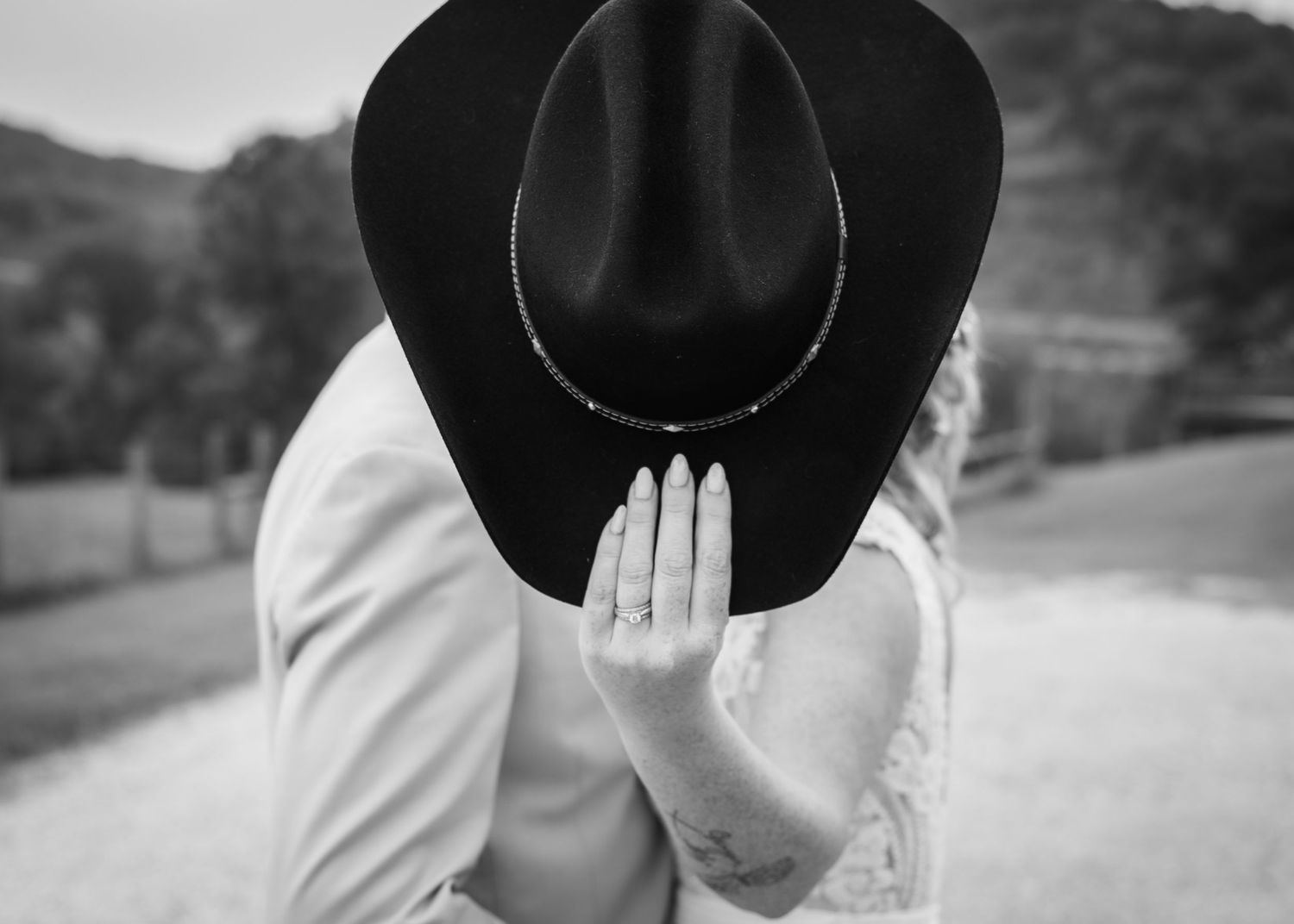 Black and white photo of a couple kissing behind a cowboy hat, showcasing an engagement ring.