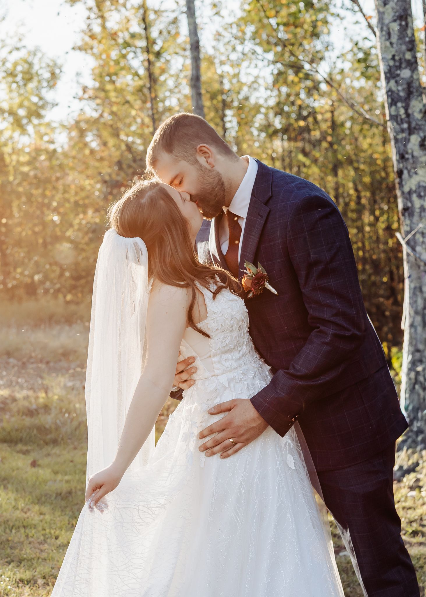 Bride and groom share romantic kiss outdoors in golden autumn sunlight among birch trees.