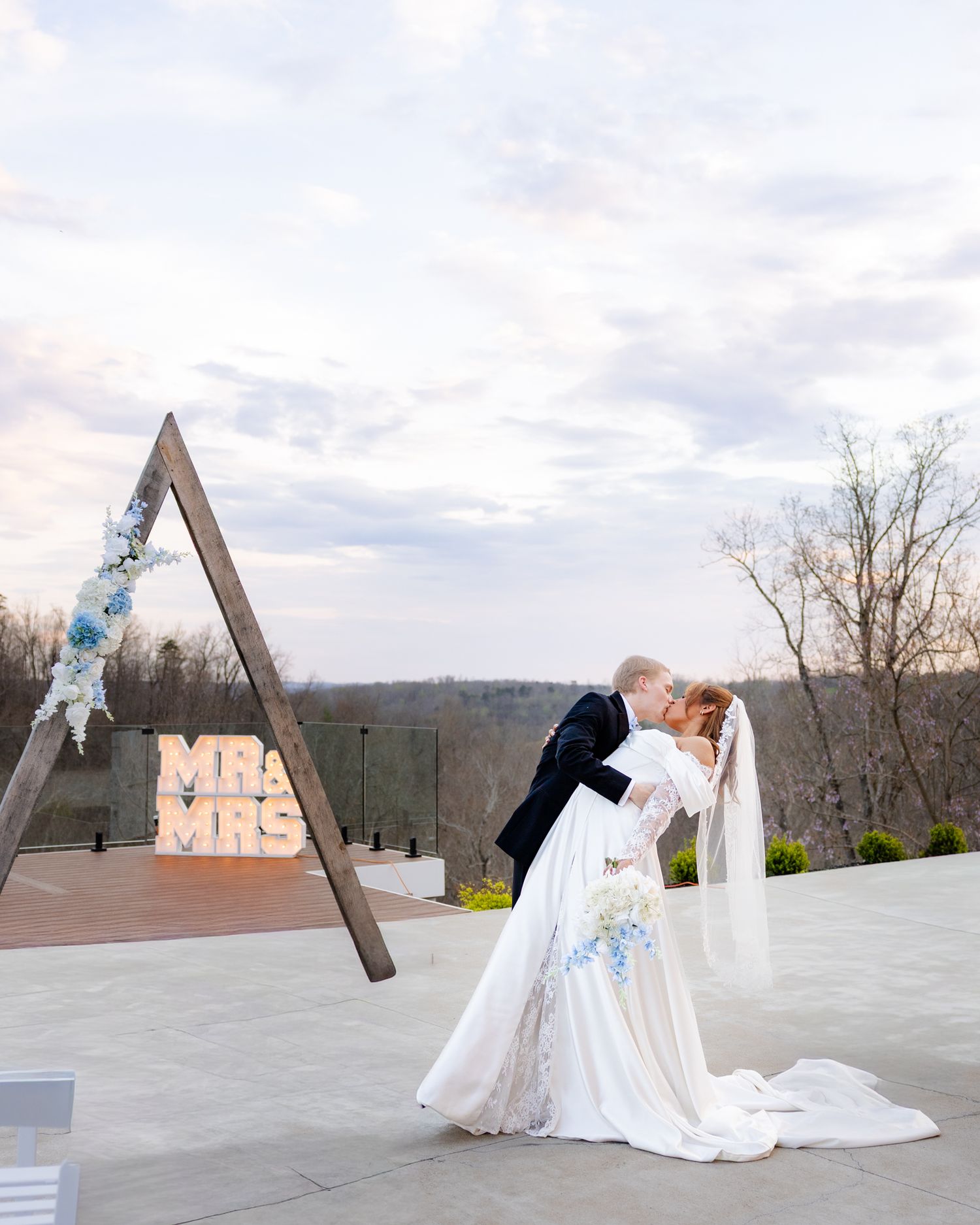 Bride and groom share a romantic dip kiss outdoors near a wooden triangle arch with lit MR & MRS marquee letters.