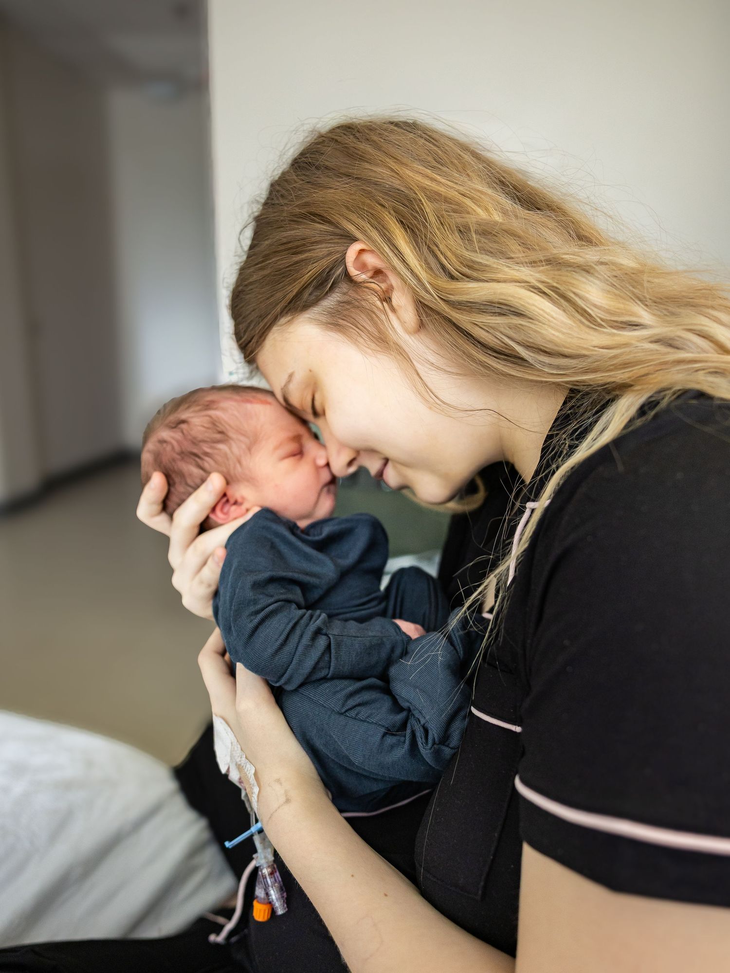 Young blonde mother tenderly touches foreheads with her newborn baby in a hospital room.