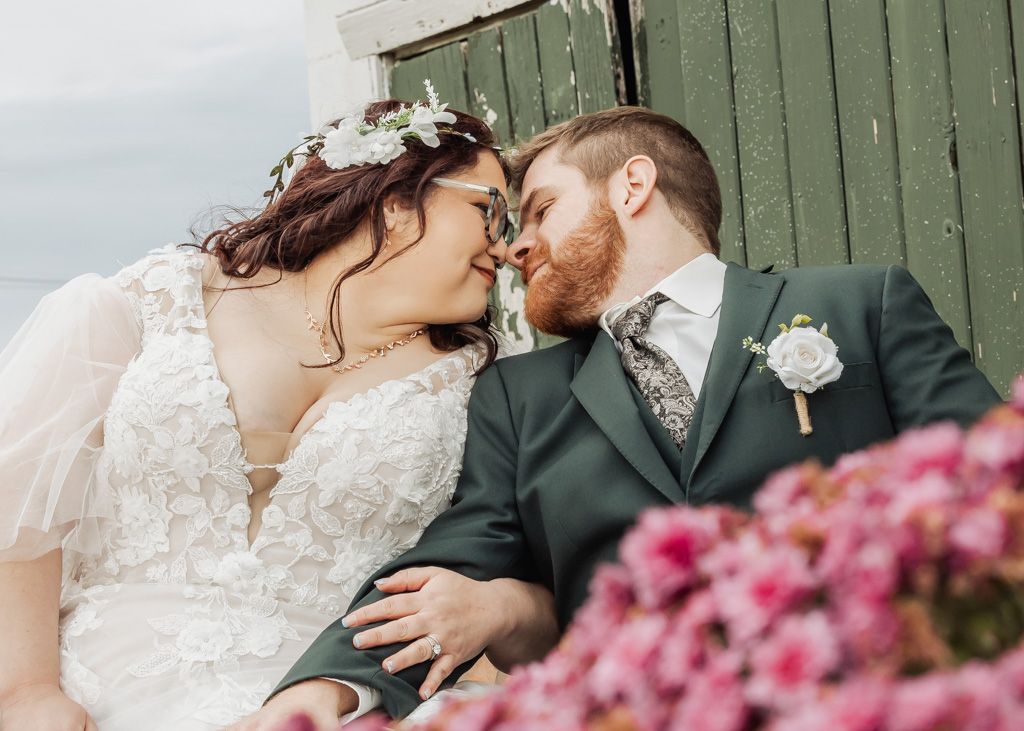 Bride and groom share a romantic moment, nearly kissing, with pink flowers and a green barn backdrop.