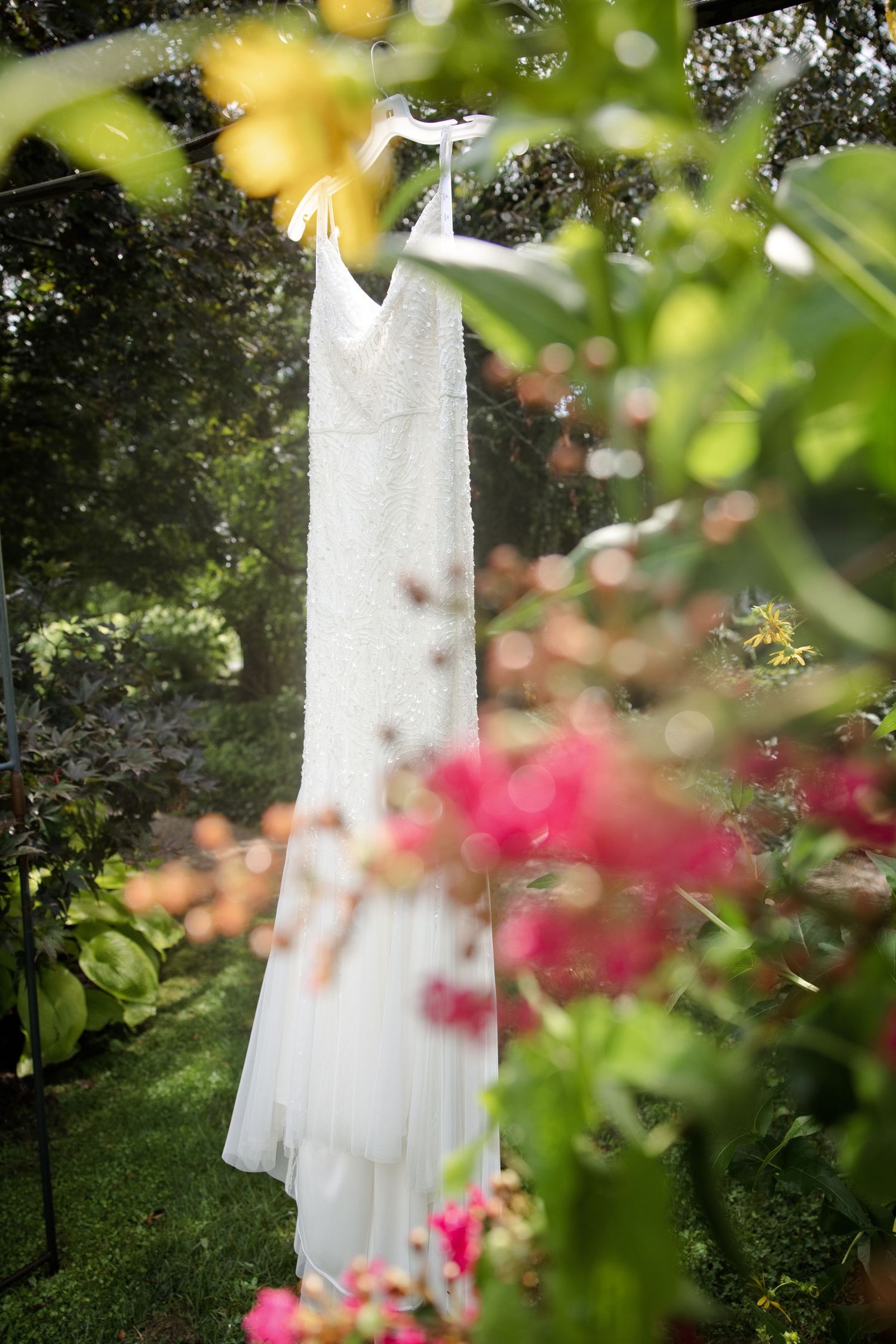 A white wedding dress hanging outdoors among lush green trees and colorful pink and yellow flowers in a garden.
