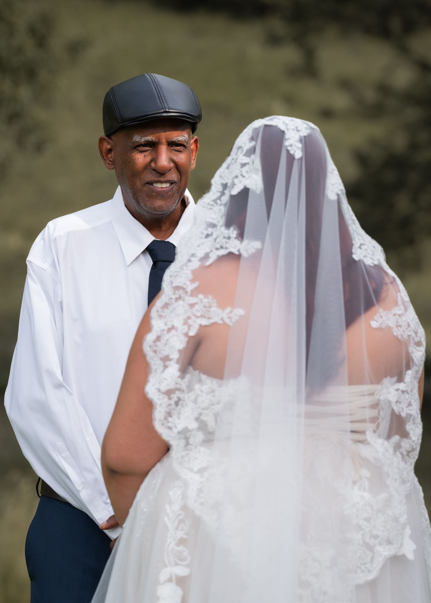 A groom in a white shirt and cap smiles beside a bride in a lace veil at an outdoor wedding.