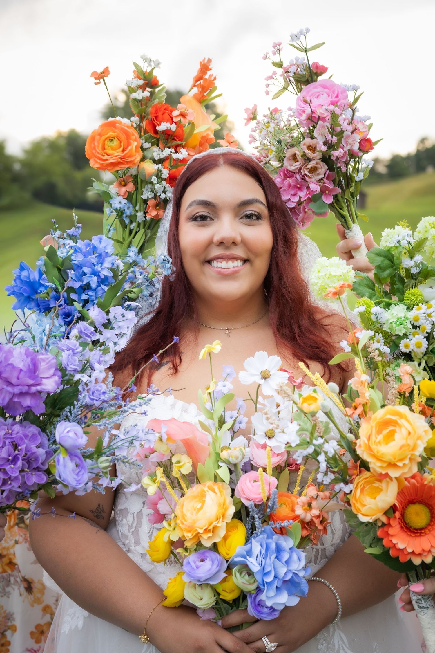 Smiling woman in white dress holding colorful floral bouquets surrounded by vibrant rainbow flowers outdoors.