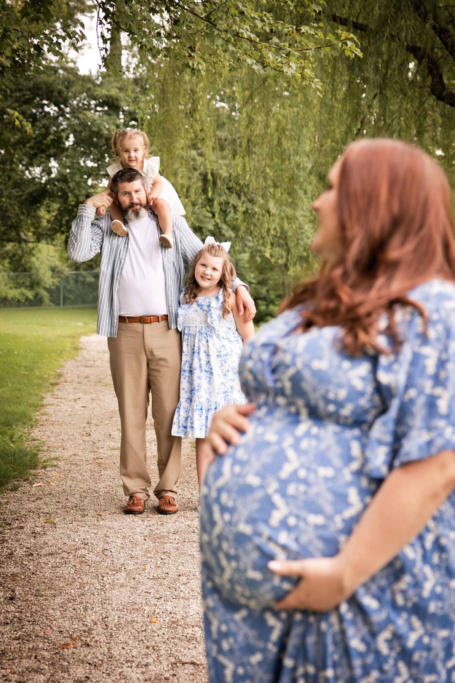 Pregnant woman in blue dress watches husband carry daughter on shoulders with another child in park.