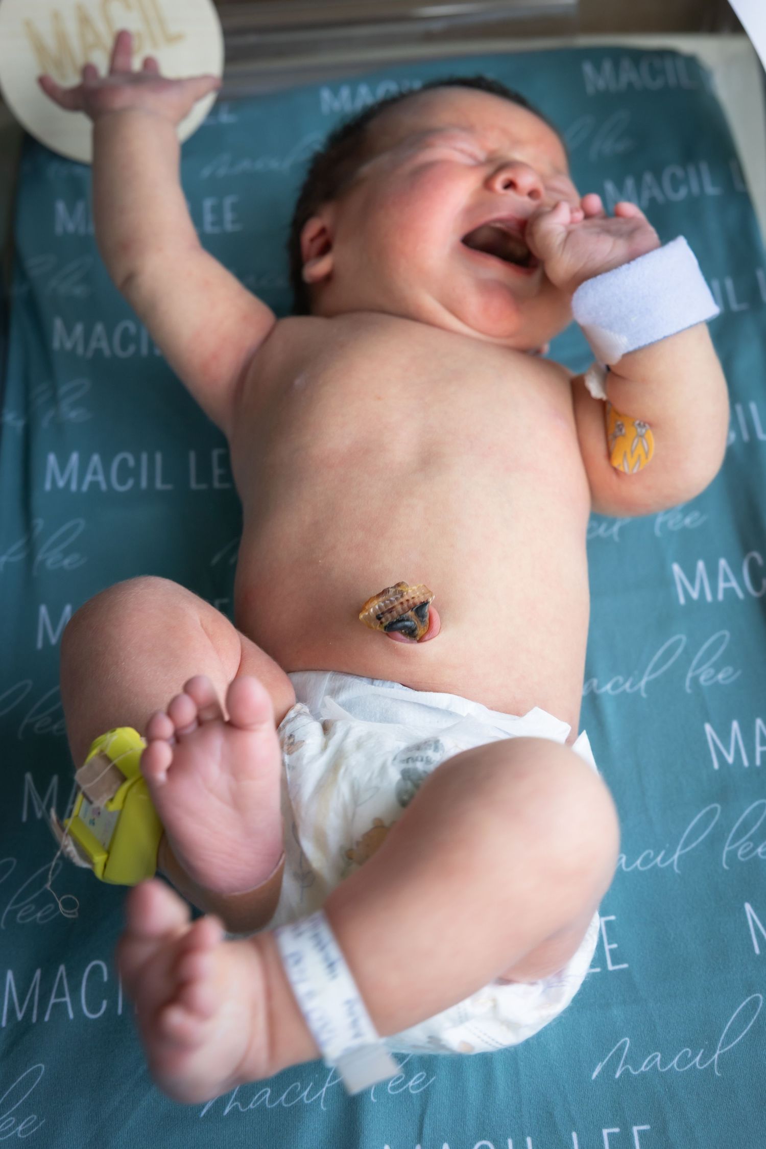 Crying newborn baby lying on a personalized Macil Lee blanket in the hospital with ID tags.