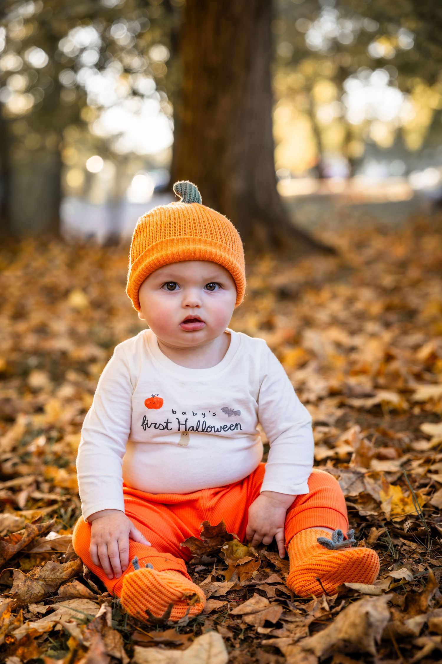 Baby in an orange pumpkin beanie and first Halloween shirt sits on fallen autumn leaves in a sunlit park.