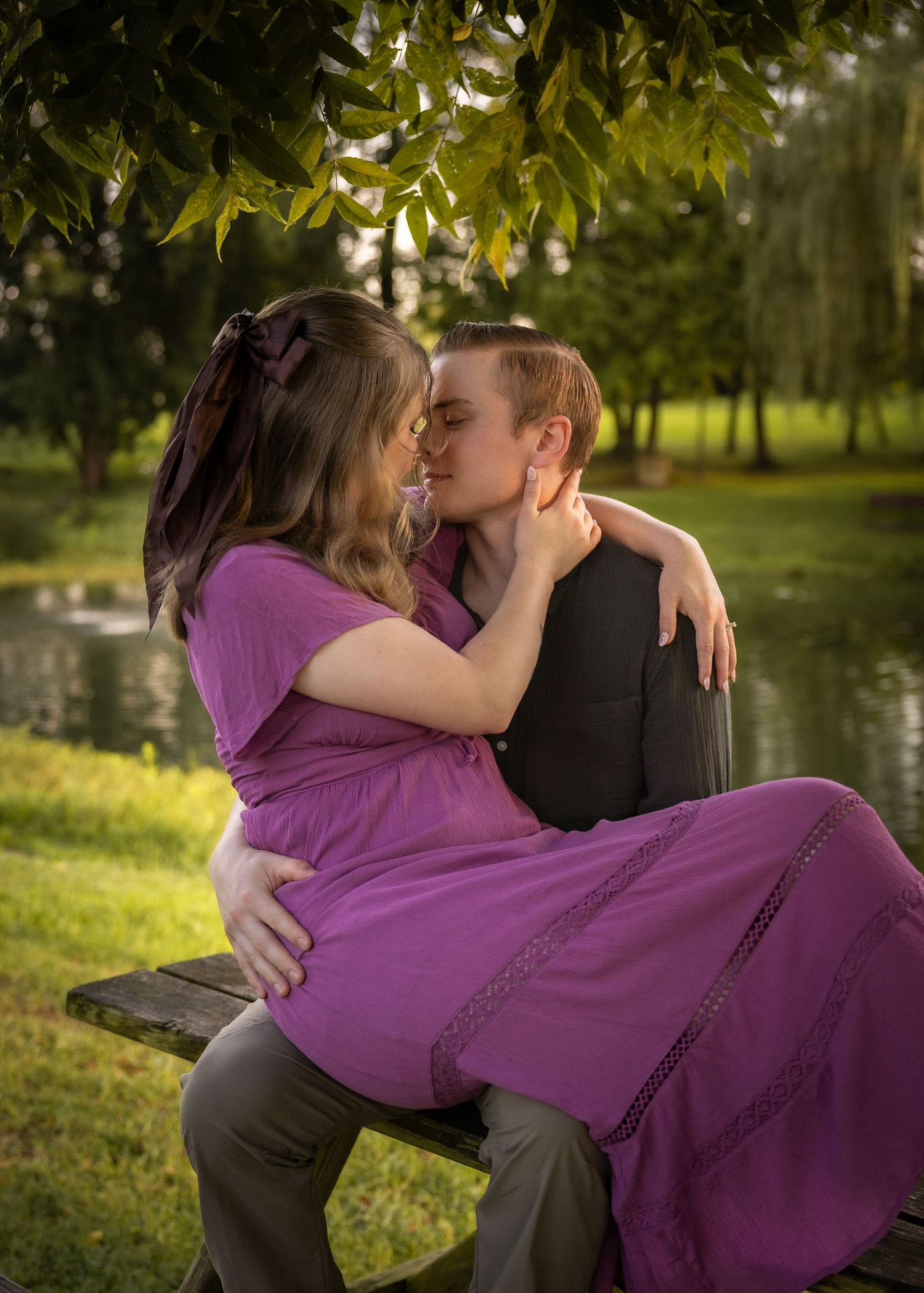 Couple sharing a romantic kiss on a park bench beside a pond, surrounded by lush green trees in warm golden light.