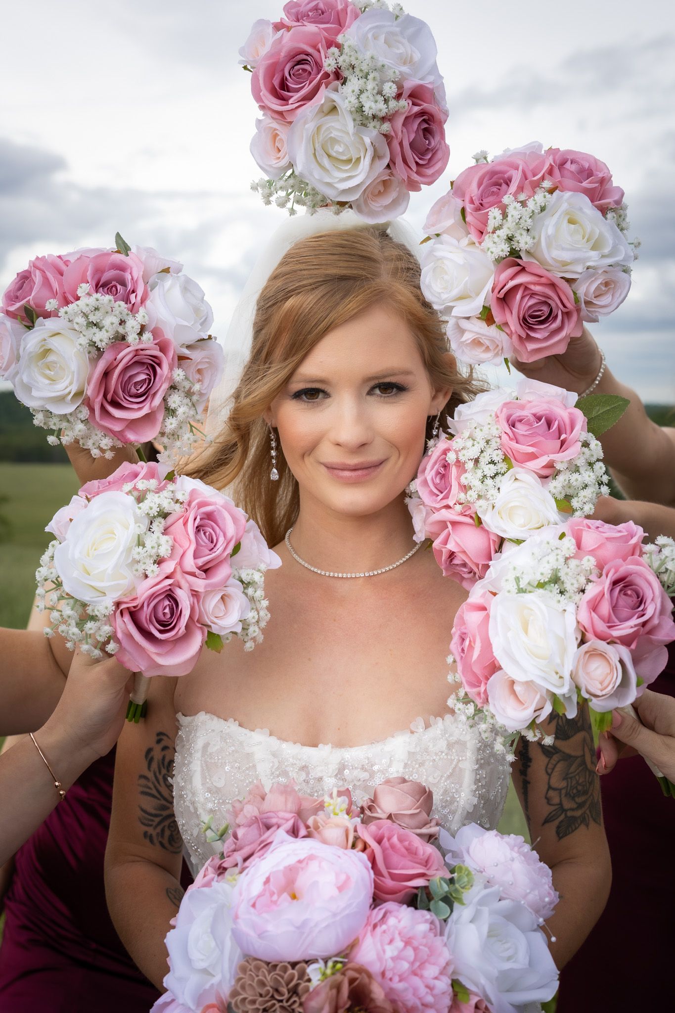 A smiling bride surrounded by bridesmaids holding matching pink and white rose bouquets outdoors.