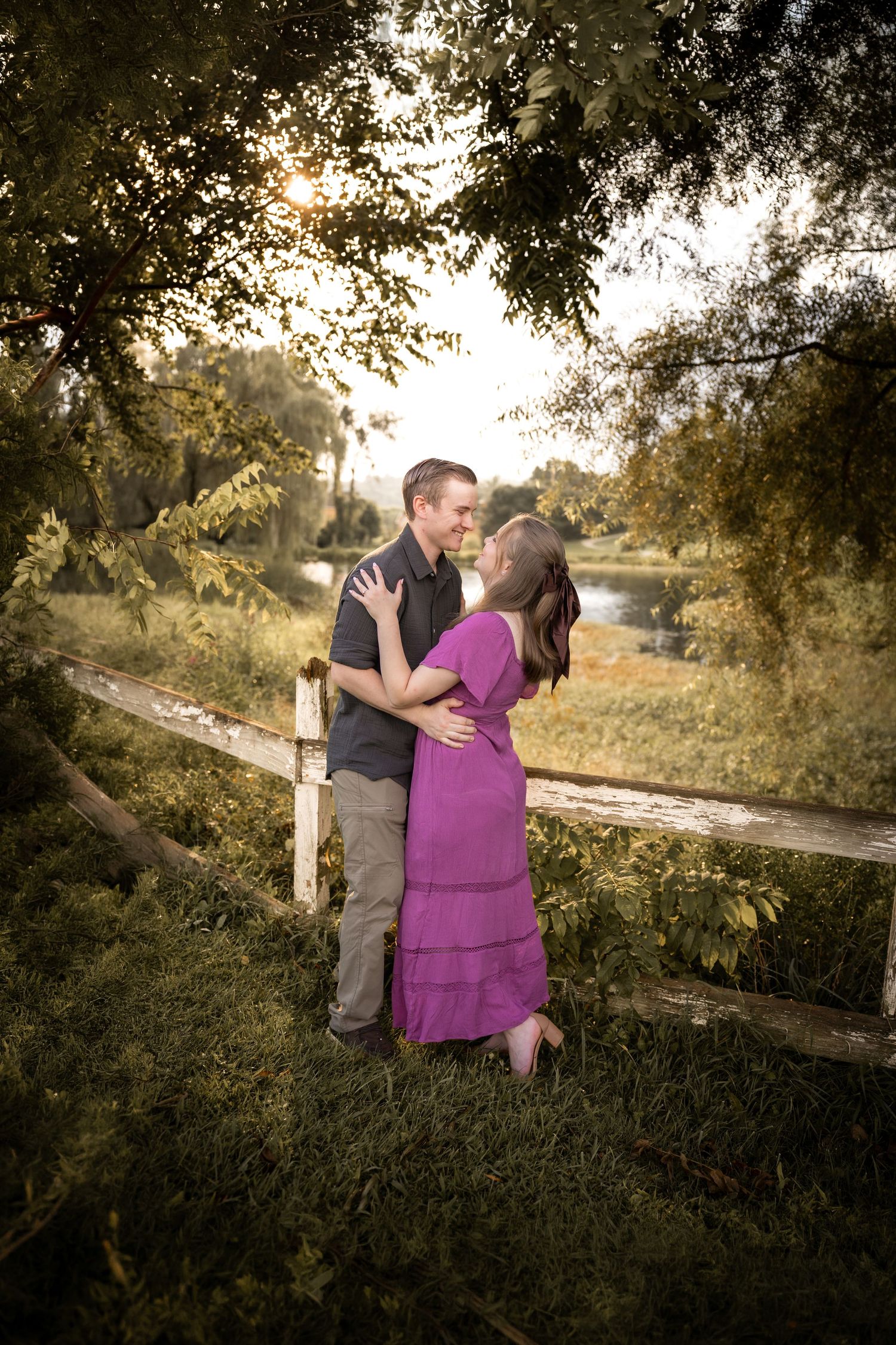 Couple sharing a romantic moment by a rustic fence surrounded by lush green trees near a serene pond at golden hour.