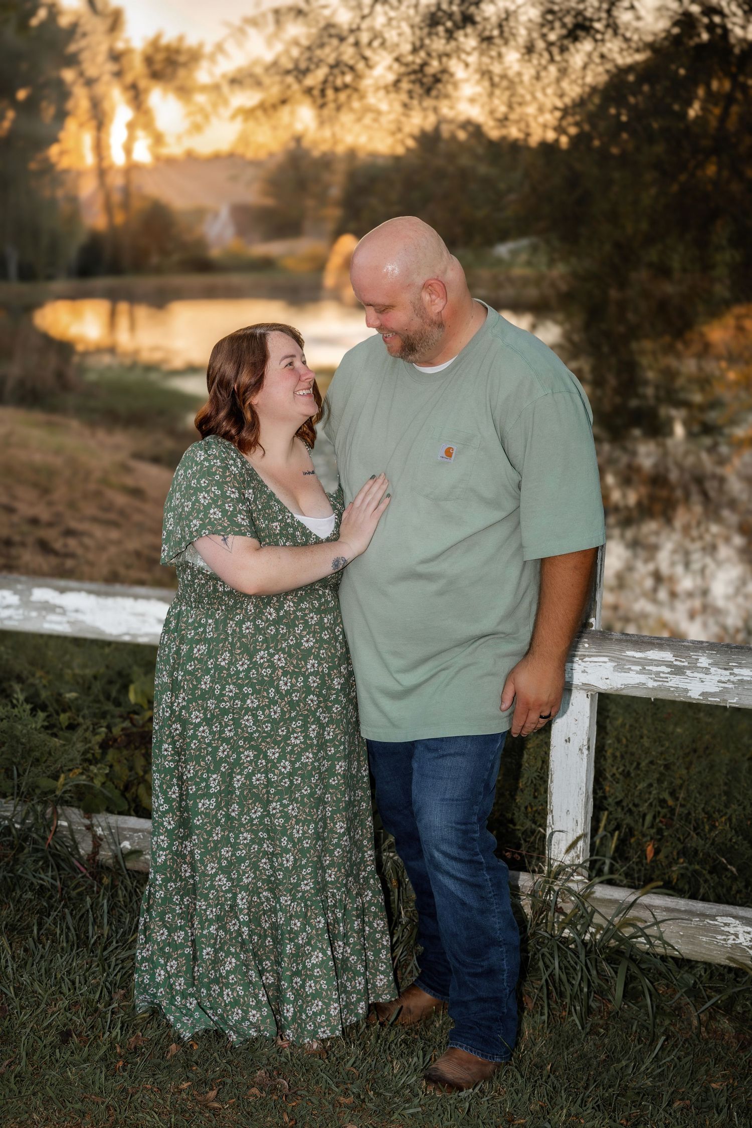 A couple shares a tender moment by a white fence at golden hour, she in a green floral dress, he in a sage t-shirt.
