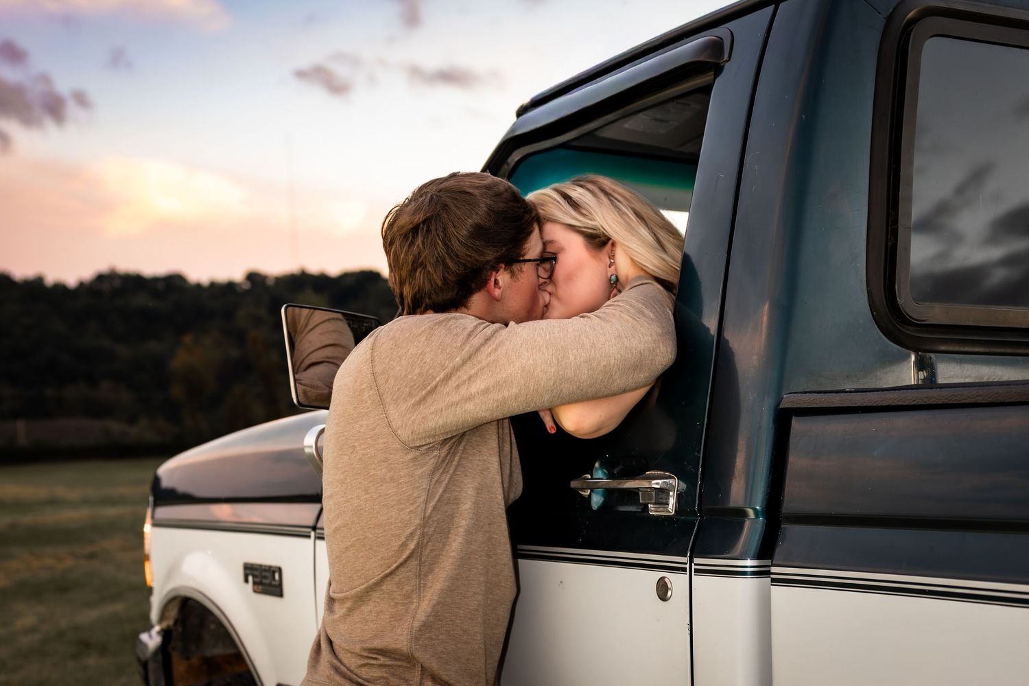 Couple sharing a romantic kiss through a truck window at sunset in a scenic outdoor setting.