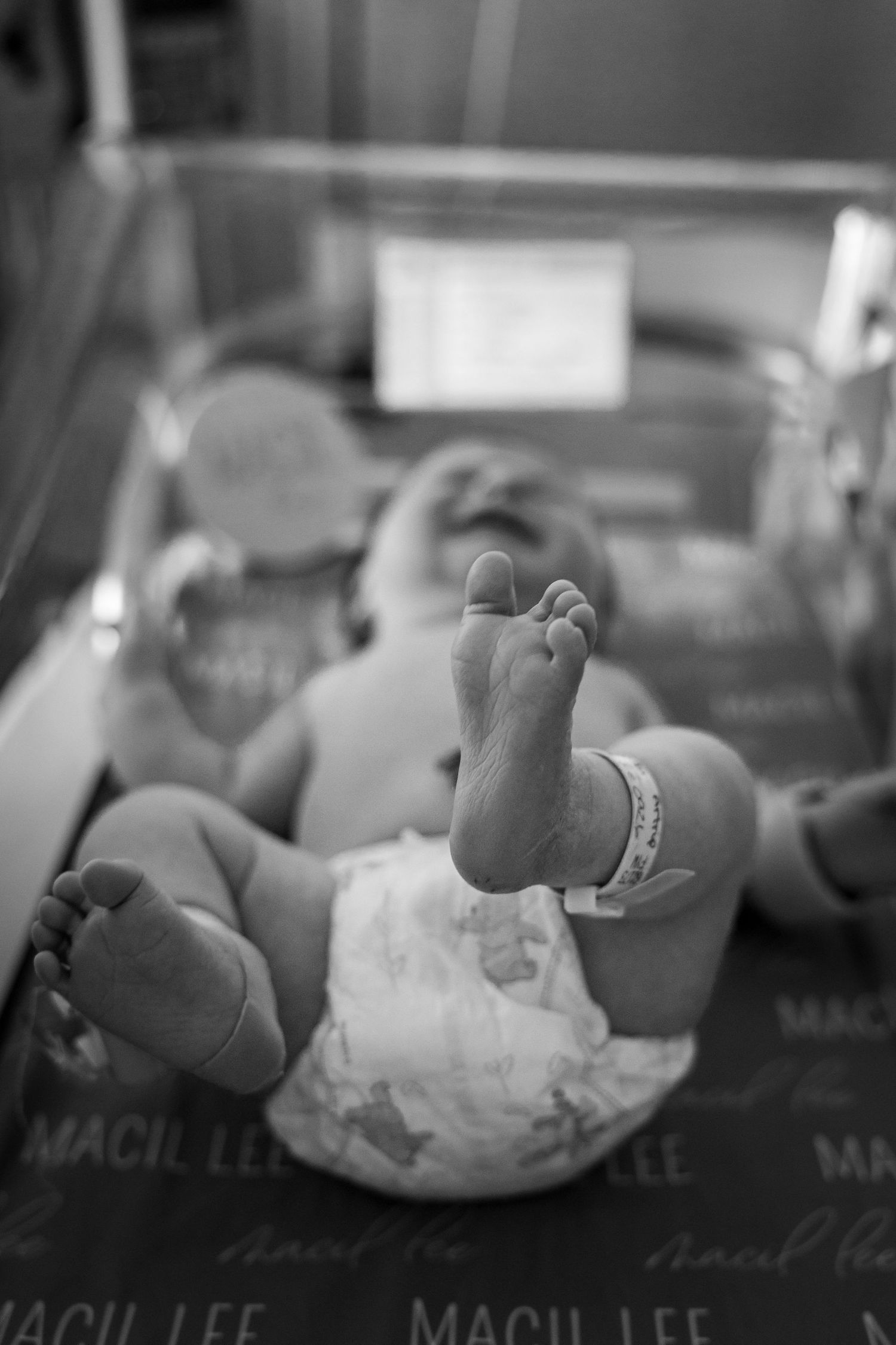 Newborn baby lying in hospital bassinet on Macil Lee blanket in black and white photo.