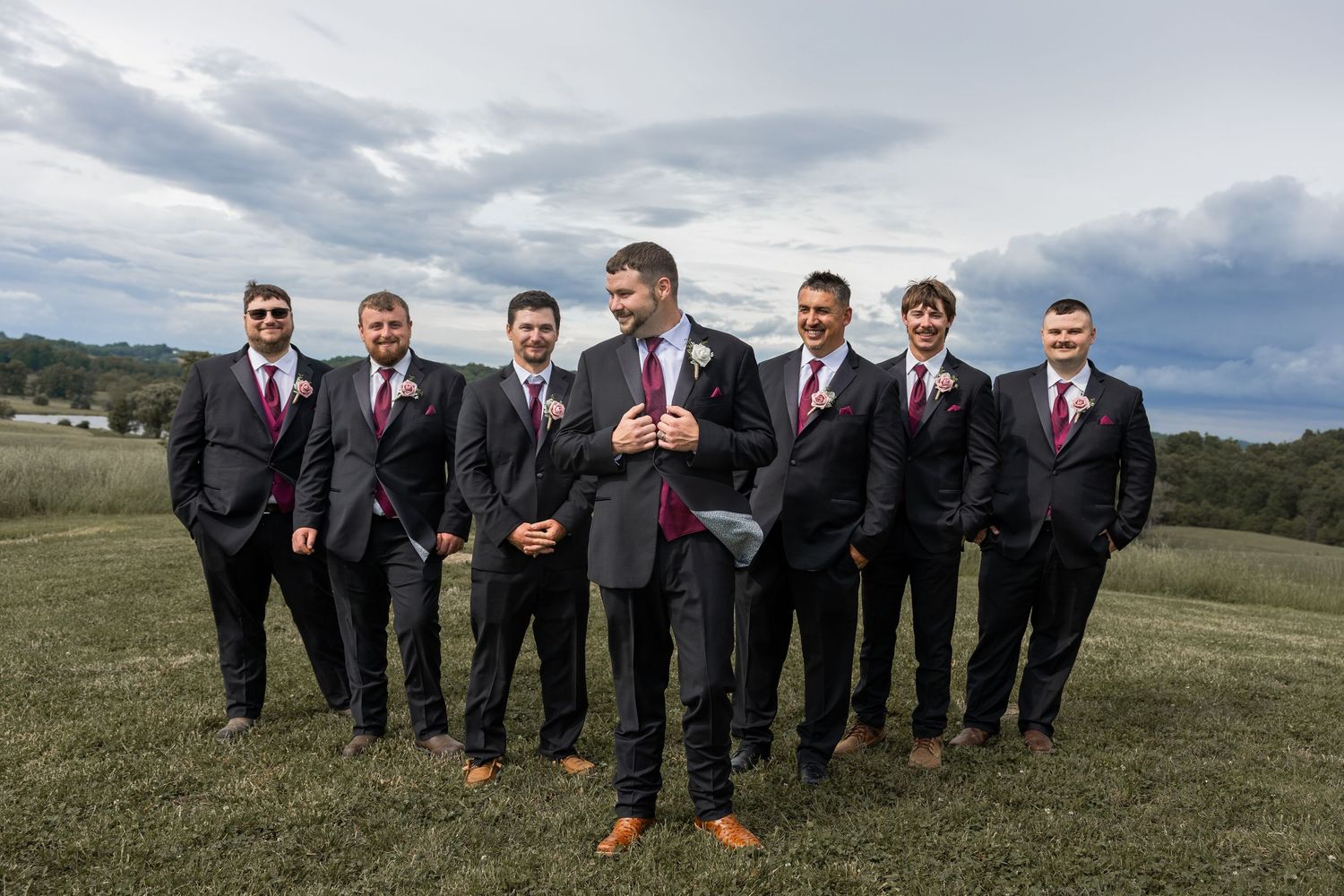 Groom stands confidently with six groomsmen in dark suits and burgundy ties on a green field under cloudy skies.