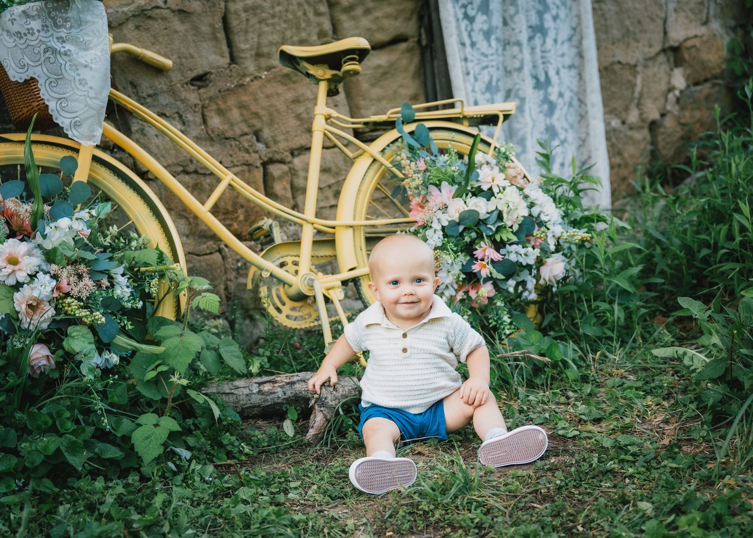 Happy baby boy sitting on grass beside yellow bicycle decorated with flowers.