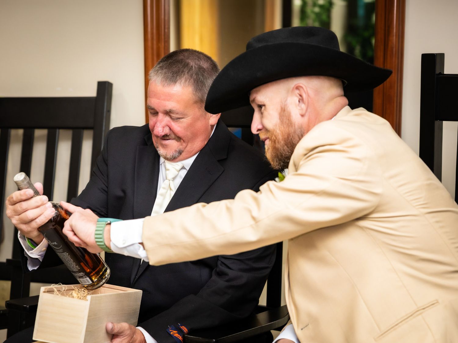 Two men in formal attire examine a bottle of whiskey, one wearing a black suit and the other a tan suit with cowboy hat.